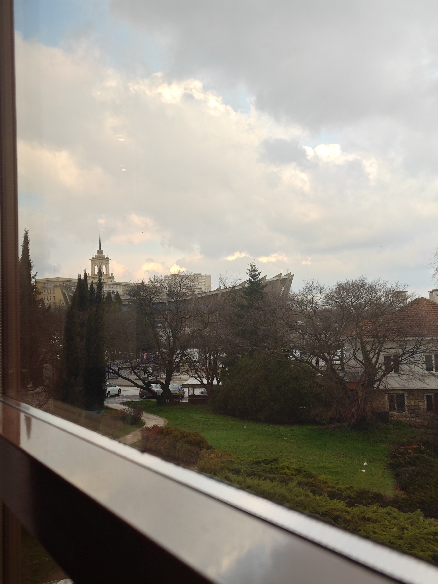 View from a window in Varna, Bulgaria, showcasing a cityscape under an overcast sky with white clouds. In the middle ground, green grass and bushes occupy the lower half of the frame, leading to a row of trees with bare branches. Further back, two distinct buildings stand out: On the left, a white building with a central tower featuring an elaborate spire, and to the right, a modern-looking building with a series of sharply angled, grey roofs. Cars parked along a road are visible between the trees. A two-story residential building with a red tile roof is nestled between the trees on the right side of the image. The foreground features a blurred window frame.