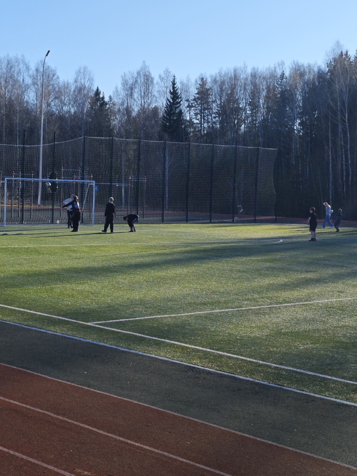 The image shows a soccer field with a running track in Markovskii, Russia. The field is artificial turf with white lines. A soccer goal is visible on the left side of the field, with a net behind it. A group of children is present on the field, some near the goal and others further away. The weather appears to be sunny. The background includes trees and a light pole. The running track is red with white lane markings.