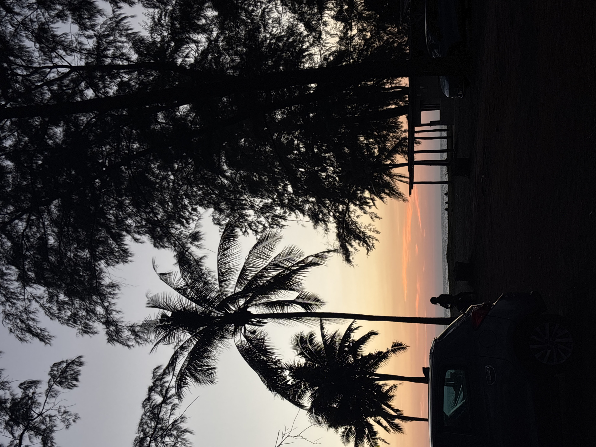 This photo was taken at Choeng Thale, Thailand. It depicts a sunset at the beach. The foreground features the silhouetted forms of tall trees including palm trees. A car is parked on the right, with a silhouetted person standing nearby. In the distance, a wooden structure stands near the water’s edge. The sky is multicolored, with orange near the horizon.