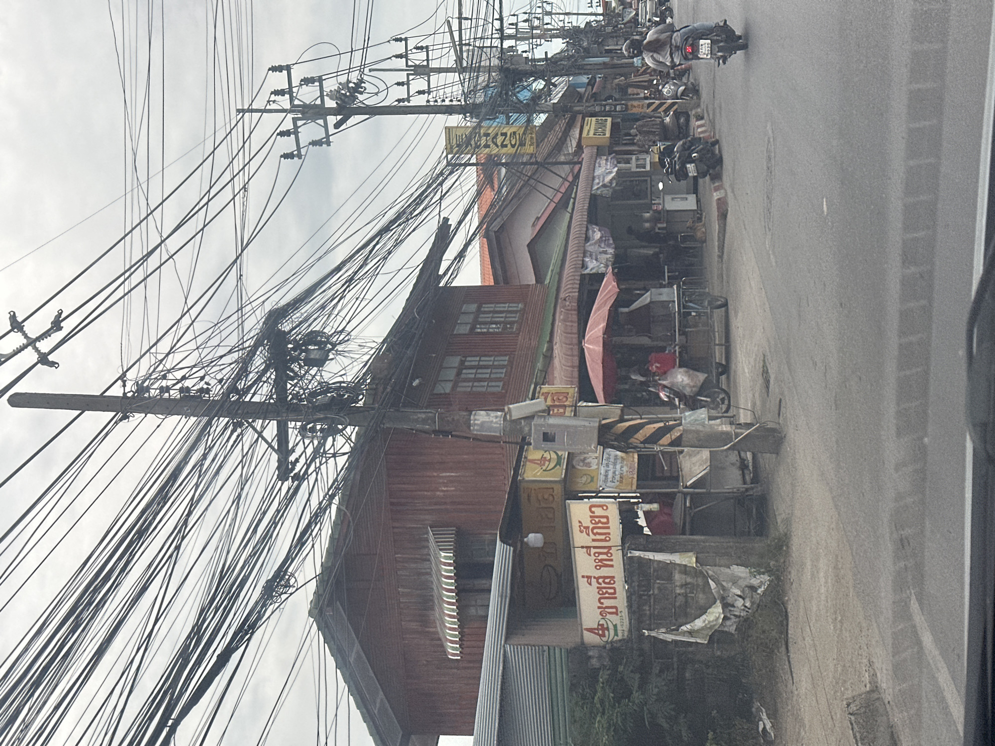 The image depicts a street scene in the Ko Samui District of Thailand, characterized by a dense overhead network of electrical wires. On the left, a two-story wooden building with a red corrugated metal exterior is visible, featuring awnings over the windows. Adjacent to it, a smaller structure with a green metal roof and a wall with graffiti-like posters is present. Several signs in Thai script are displayed, including one that reads "ชายสี่ หมี่เกี้ยว," indicating a noodle shop.
The street itself is paved, with a sidewalk lined with businesses. To the right, there are more buildings, some with orange roofs, and a yellow sign with the word "EXCHANGE" is prominently displayed. Several people are visible on foot and on motorcycles. One woman on a scooter is in the foreground, traveling away from the camera. Various stalls and shops are open, some with red umbrellas providing shade. A small dog is seen near a stall. The environment suggests a bustling commercial area on an overcast day.