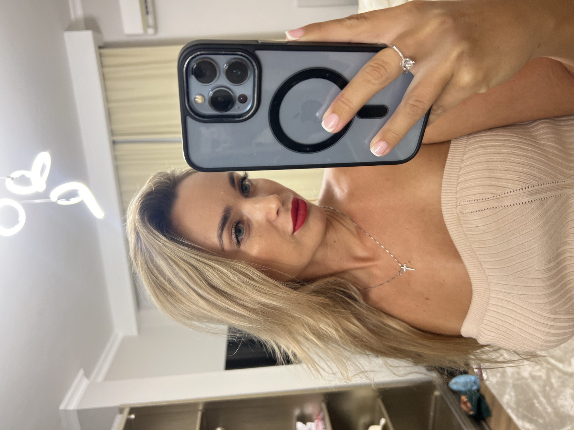 A woman is taking a selfie in what appears to be a bedroom or dressing area, with lighting fixtures visible on the ceiling. She has blonde hair, is wearing bright red lipstick, and has a cross necklace. She is holding up an iPhone in a grey case, and a ring is visible on her finger. The background shows a closet or wardrobe area with shelves, and curtains. The overall setting suggests an indoor, possibly domestic, environment. There is no indication of Sharm El Sheikh, Egypt.