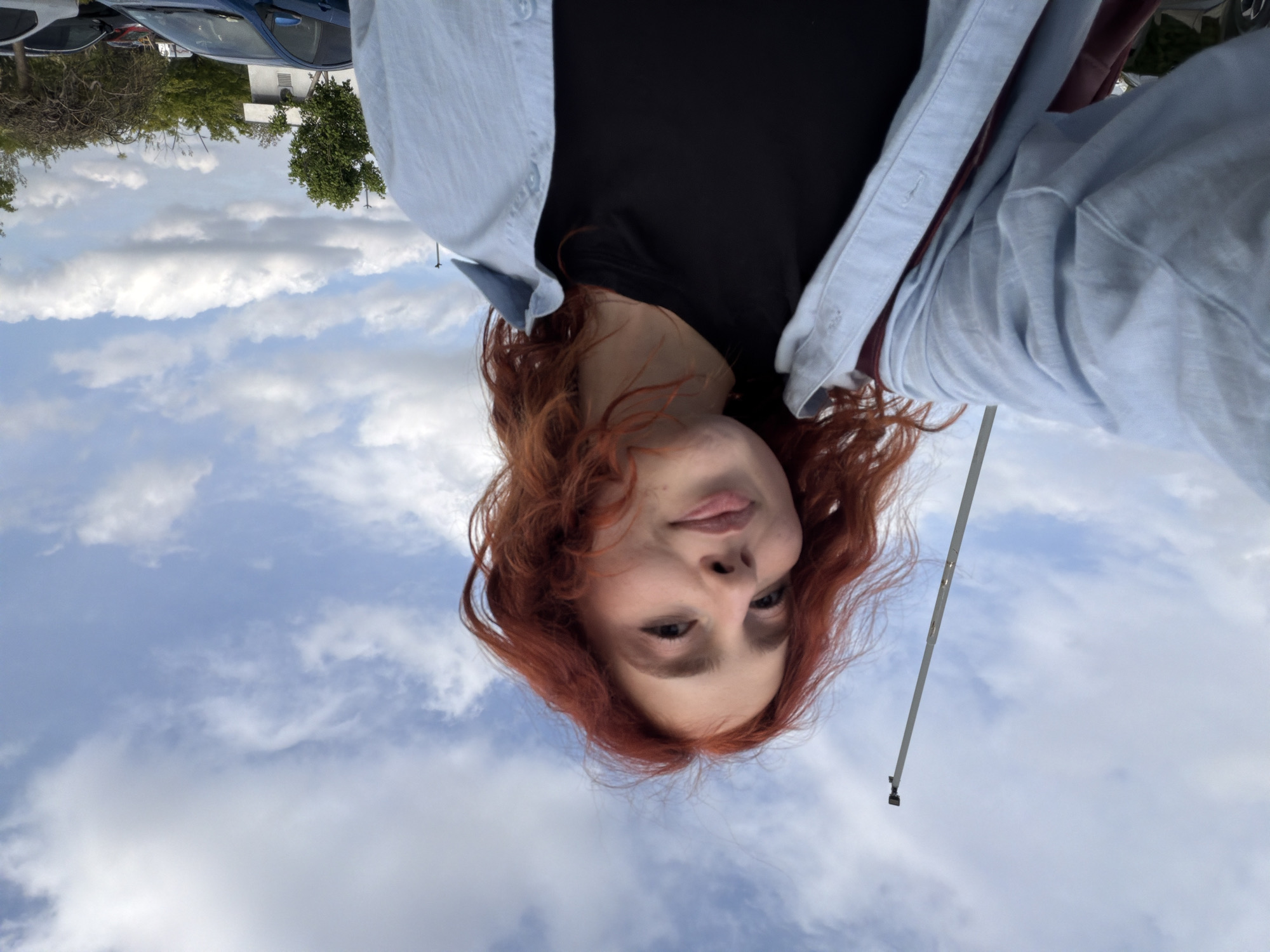 An upside-down, low-angle selfie captures a young person with reddish-brown, wavy hair looking directly at the camera. They wear a black t-shirt beneath an unbuttoned light blue collared shirt. Their expression is neutral, with a slight hint of a smile or smirk.
The background is dominated by a bright blue sky filled with scattered white clouds, suggesting a clear daytime setting. Visible at the top of the frame (when viewed correctly) are the roofs of several cars, implying a parking area or street scene. Beyond the cars, green trees and some distant buildings are discernible, indicating an urban or suburban environment in Tbilisi, Georgia. A thin, light-colored pole, possibly a selfie stick or a light fixture, extends into the frame on the right side. There is no visible text in the image.