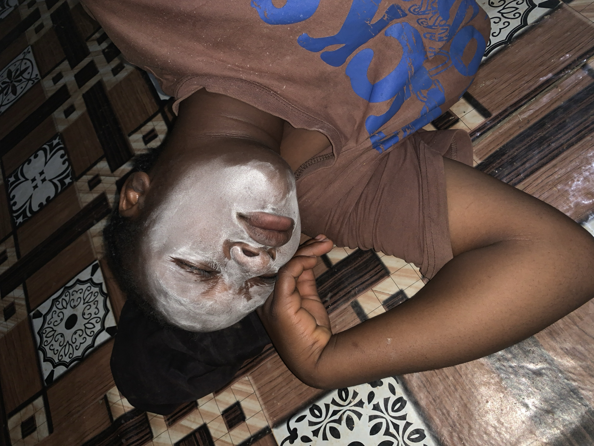 A person lies on a tiled floor, presumed to be indoors, in Agban, Nigeria. The floor tiles are a mix of brown, black, and white patterns. The person wears a brown t-shirt with a blue design and is lying on their back with their head turned to the left. Their face is covered in a white substance, possibly a facial mask. Their right arm is bent and resting near their face. The overall impression is of someone relaxing or undergoing a skincare treatment.