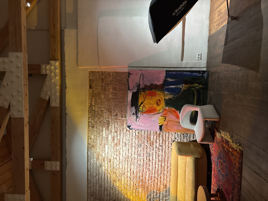 This image captures an interior space with an artistic and industrial aesthetic, possibly a studio or a loft. The perspective is from an elevated position, looking down slightly, and the image is oriented vertically (as if a phone was held in portrait mode and the scene is wider than tall).
On the far left, a significant structural element dominates, consisting of large, light-colored wooden beams arranged vertically and horizontally, connected by white metal plates with visible rivets. These form a sturdy, open framework.
Moving to the right, there's a light-colored wall, part of which transitions into an exposed red brick wall section. A prominent feature on the brick wall, spanning across the light-colored wall above it, is a large, colorful abstract painting. The painting depicts a stylized figure with a yellow-orange face, large eyes, and a black hat, set against a background of pink, blue, and dark green shapes. The brushstrokes are loose and expressive.
Above the painting, on the white wall, a black professional photography softbox, labeled "Profoto Softbox RFi", is visible, attached to a stand that extends upwards out of frame. Below the softbox, on the white wall, a standard electrical outlet is visible. A faint, reddish-orange laser line stretches horizontally across the white wall, just above the brick section and the painting.
In the bottom right portion of the image, two pieces of furniture are present. A plush, ribbed, mustard-yellow sofa or armchair is seen, partially cut off by the bottom edge. To its right, a uniquely shaped, abstract white chair with a sculptural, curved form stands. Further to the right and slightly behind these chairs, a colorful patterned rug, predominantly red with blue and white designs and fringed edges, lies on a dark wooden floor.
The lighting in the room appears to be mixed. There's a warm, yellow light illuminating the brick wall and the front of the yellow sofa, creating a cozy glow. Other parts of the room are in shadow or lit by ambient light, giving the scene a dynamic and inviting atmosphere.