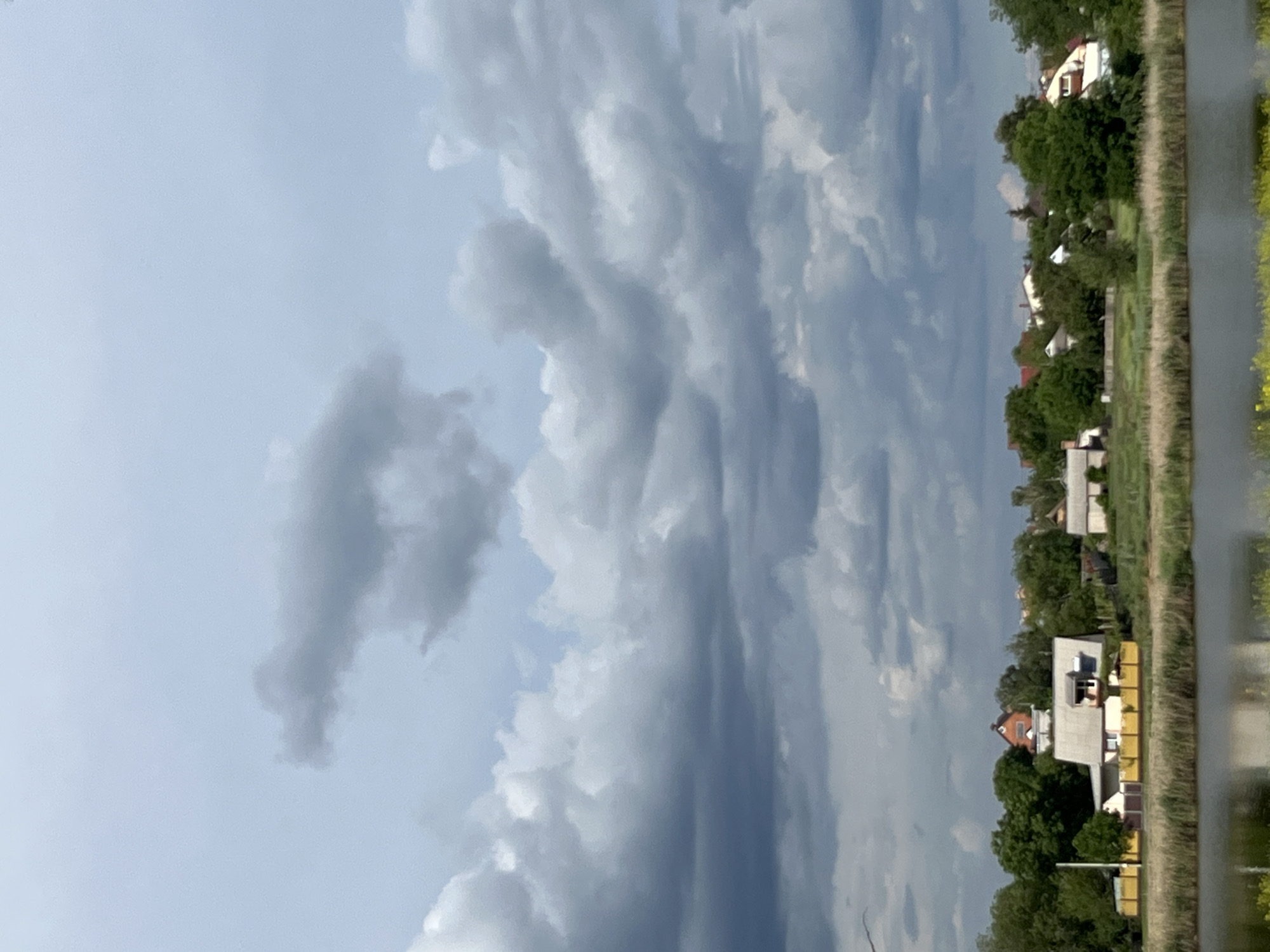 A low-angle, medium shot captures a cloudy sky over a suburban landscape adjacent to a body of water. The sky is filled with various cloud formations, ranging from light, wispy clouds to darker, more ominous storm clouds. In the upper left quadrant of the sky, a peculiar cloud formation resembles a stylized, elongated animal with a distinct head and tail.
Below the sky, a line of trees and houses is visible along the edge of a calm body of water in the foreground. The houses are modest, with pitched roofs and some exhibiting a yellow fence in front. The vegetation is lush and green, with tall grasses growing along the water's edge. The overall atmosphere suggests a somewhat overcast day in a rural or suburban area.
This image was taken on July 10, 2023, at 10:45 AM in Kryvyi Rih, Ukraine.