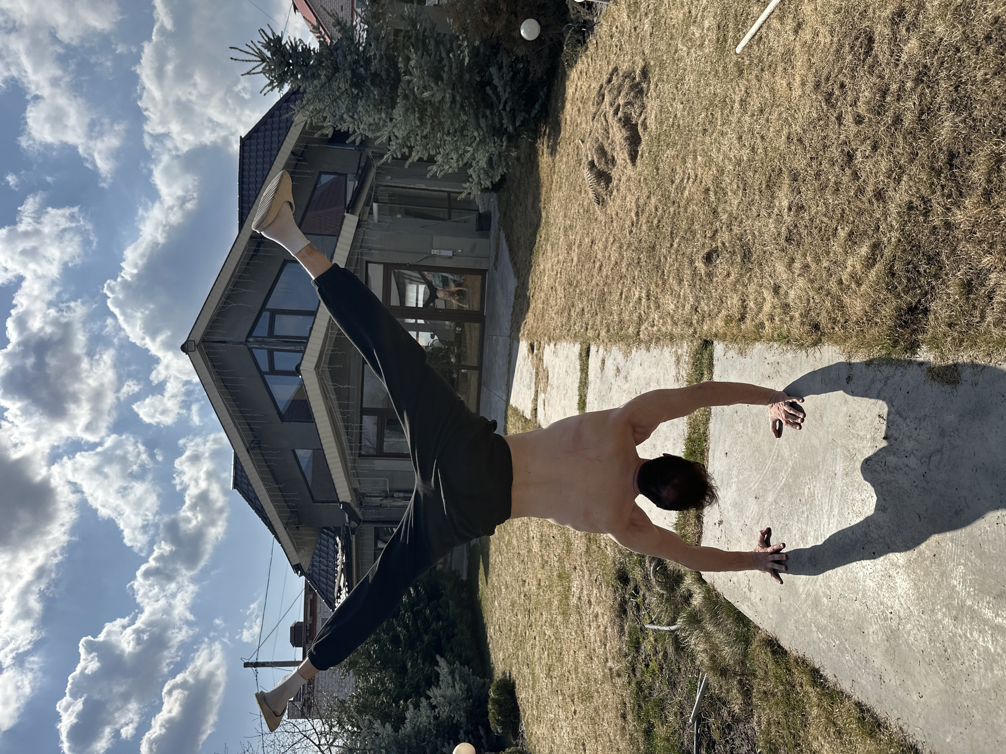A man is performing a handstand split outdoors on a concrete path. He is shirtless and wearing dark-colored pants with light-colored shoes. The man's head is closest to the concrete path. His legs are split in a wide angle, with one leg pointing towards the sky and the other towards the ground. In the background, there is a two-story building, grass, and a blue sky with clouds. The photograph appears to have been taken during the day. There is no reliable information in the image that would confirm that it was taken in Kursk, Russia.
