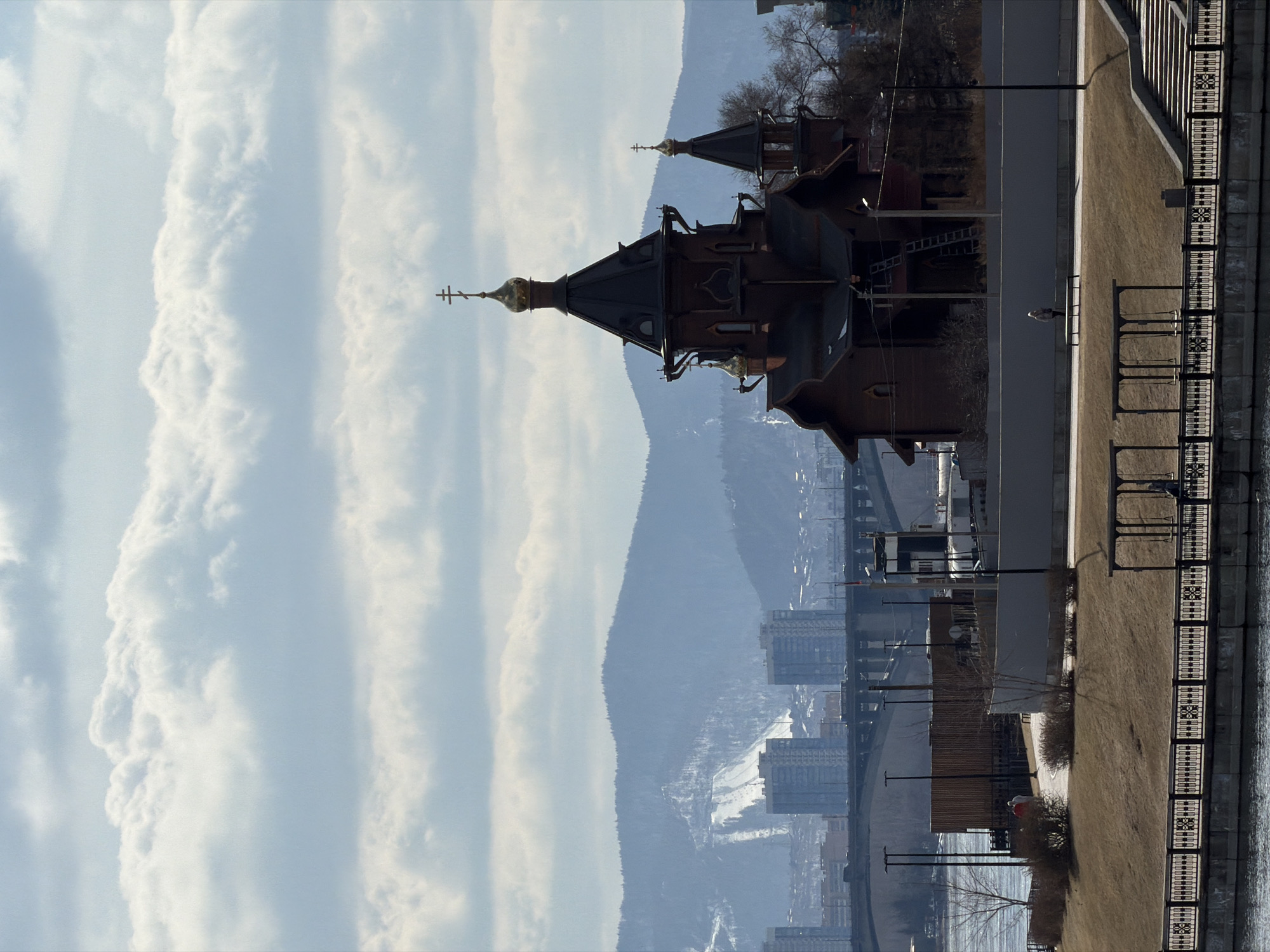A panoramic, rotated daytime image captures a city scene from a high vantage point, featuring a prominent church, distant mountains, and a waterfront area.
On the left, a large, dark-hued Russian Orthodox church stands out, characterized by its distinctive dome-shaped spires topped with golden crosses. The main dome is intricately designed, and a smaller dome is visible behind it, showcasing traditional architecture.
In the background, hazy, blue-tinged mountains rise, possibly with snow-covered patches, forming a rugged natural backdrop. Several modern, multi-story urban buildings are scattered across the mid-ground, nestled between the church and the mountains, indicating a developed cityscape.
To the right, a paved embankment or promenade runs alongside what appears to be a river or large body of water. Decorative black metal railings with geometric patterns line the water's edge. Beyond the railings, a patch of brownish, dry grass is visible, interspersed with bare trees and several street lamps. A road runs parallel to the embankment. One person is distantly visible on the embankment, standing near a bench.
The scene is illuminated by natural daylight, under a partly cloudy sky with prominent, streaky white cloud formations. The bare trees and brown grass suggest a season of late autumn, winter, or early spring. No specific activities or events are actively taking place beyond the presence of the solitary figure. The distinctive church architecture and the mountainous, riverine urban setting are strong location cues for Krasnoyarsk, Russia. There is no visible text in the image.