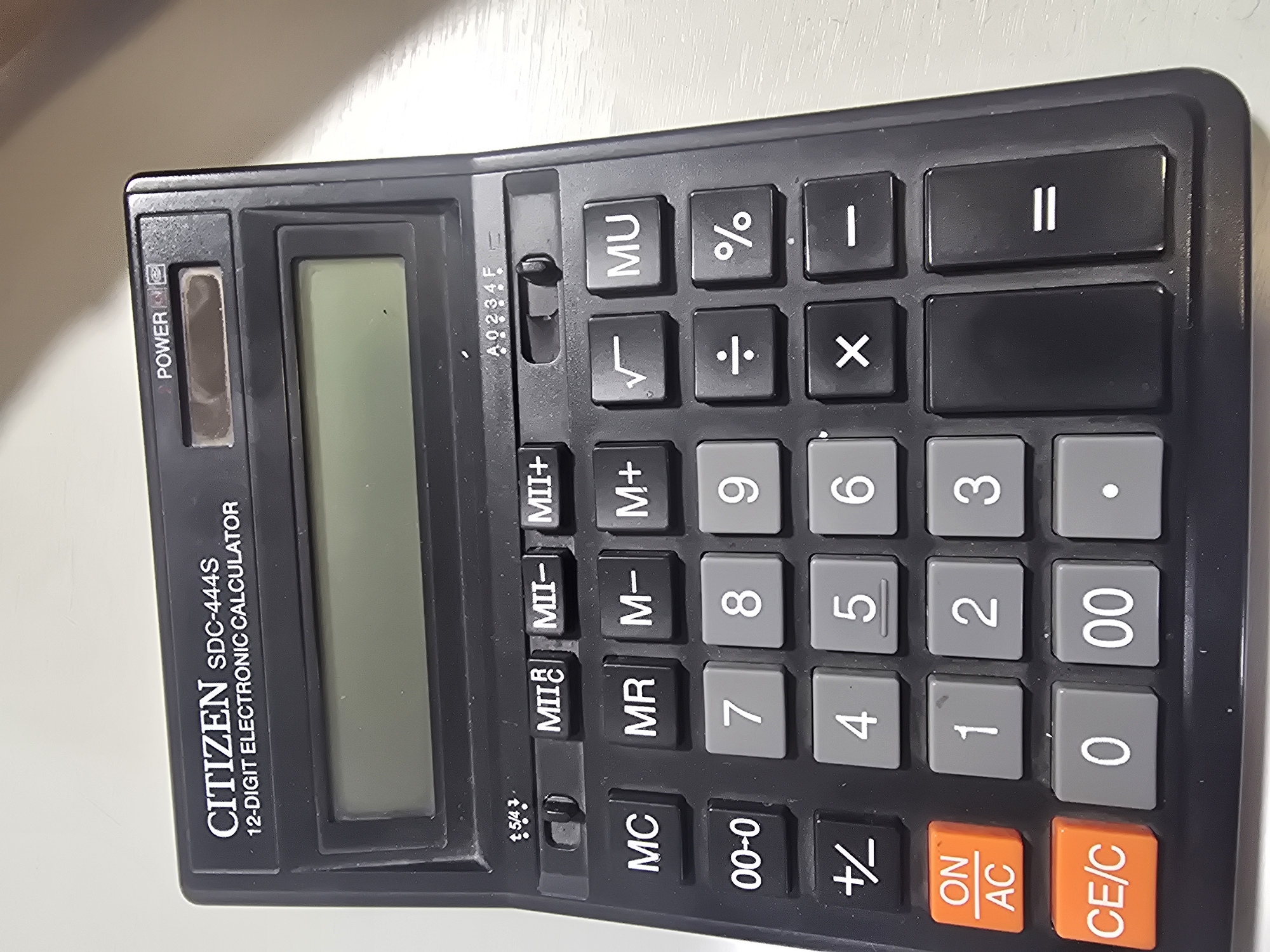 The image shows a close-up, overhead view of a black CITIZEN SDC-444S 12-digit electronic calculator. The calculator has a large digital display screen at the top, a power button, and various function and number keys. The keys include memory functions (M+, M-, MR, MC), arithmetic operations (+, -, x, ÷, =, .), number keys (0-9, 00), and special function keys (√, %, +/-). The calculator is positioned on a light-colored, textured surface, possibly a desk or table. The lighting appears to be from above and to the side, creating some glare on the screen and keys. There is no visible indication of the calculator's location within the city of Ulan-Ude, Russia; the image solely focuses on the object itself.