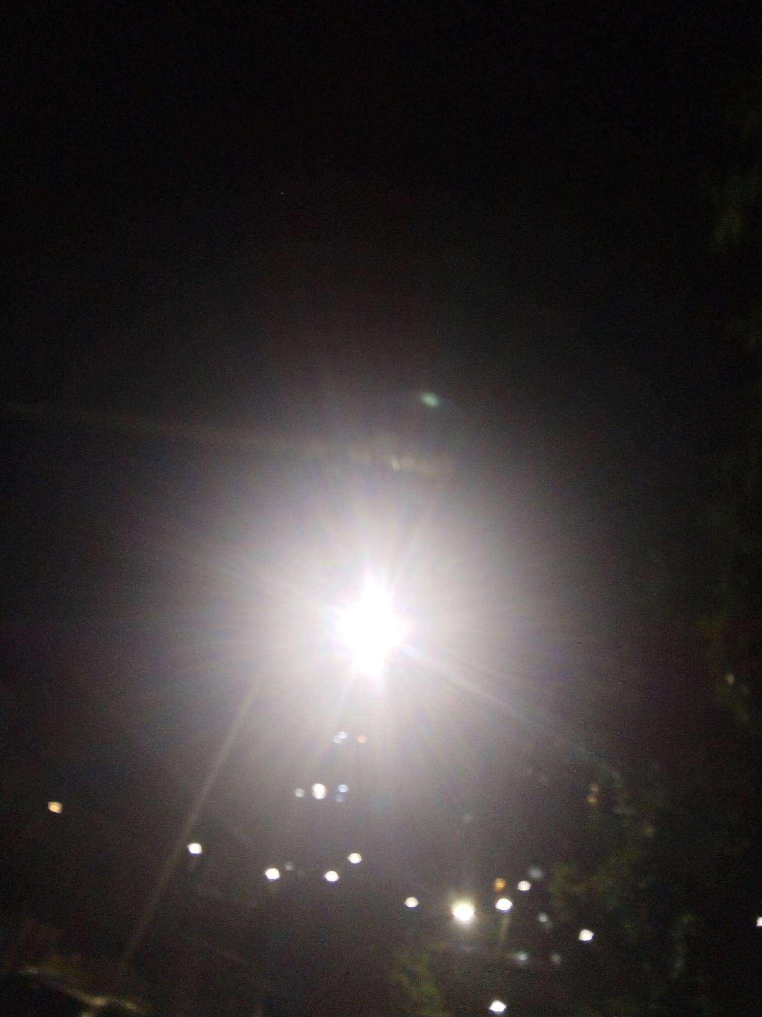 The photograph was taken at night in what appears to be Jiulongpo District, China, as indicated by the streetlights and possible presence of trees on the right. The primary subject is a bright light source, likely a streetlight, positioned centrally and dominating the frame. The light is so intense that it creates significant lens flare and overexposure, obscuring details. The foreground and background are predominantly dark, with scattered points of light suggesting other streetlights or distant building lights. The image is slightly out of focus.