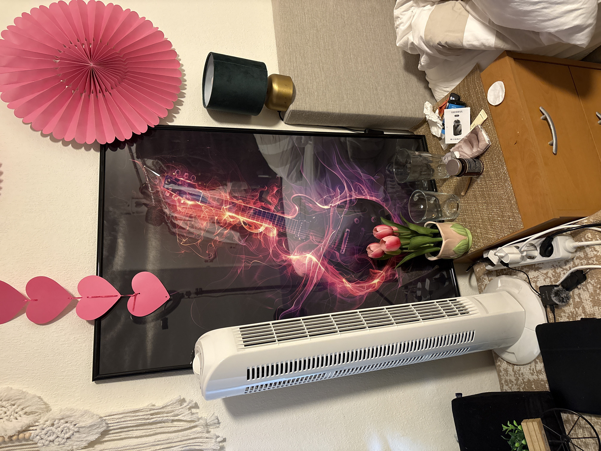 The image shows a cluttered bedroom nightstand next to a bed in Heerlen, Netherlands. On the nightstand, there is a white tower fan, a framed poster of a guitar with abstract fiery elements, a pink decorative paper fan, a small potted plant with pink tulips, and two empty glasses. There is also a small green lamp with a gold base sitting on the headboard of the bed. The nightstand surface is covered with various items including a bottle of medication, a small bag, and a makeup wipe. A power strip with several plugs is connected to the fan and other devices. To the left of the nightstand is a black case for a tablet. A macrame wall hanging is visible on the wall to the left of the fan, and a string of pink heart cutouts hangs above the framed poster. The bed on the right is unmade and covered with white bedding.
