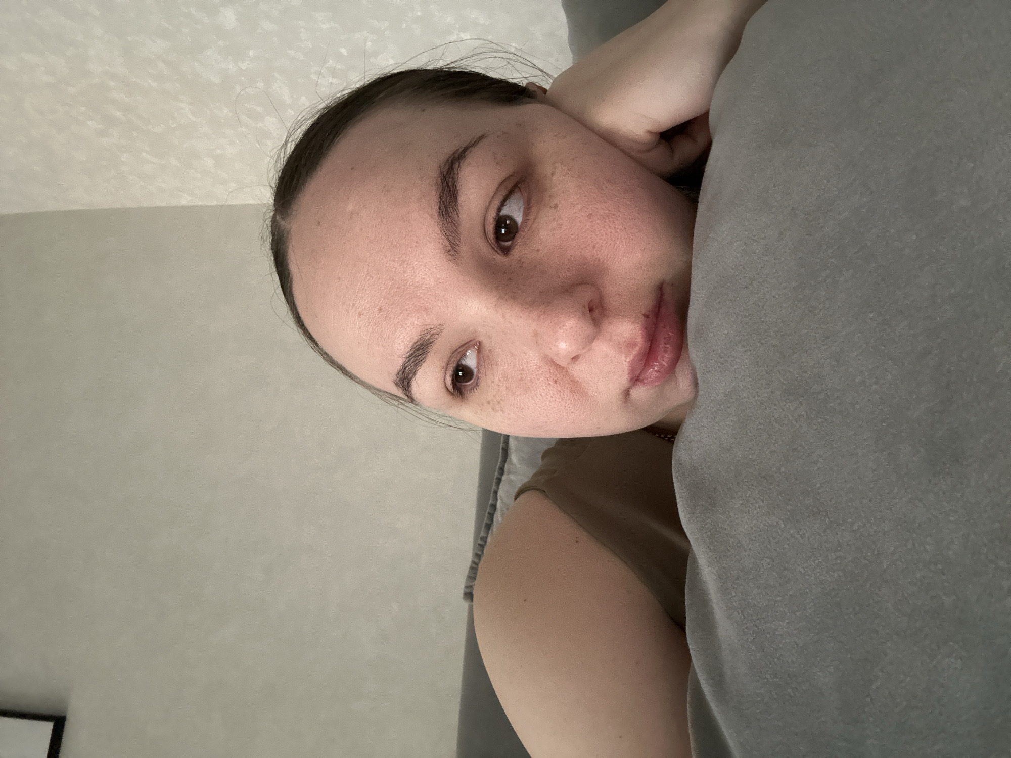 A young woman is lying down, resting her head on a gray pillow. She has brown hair pulled back, fair skin with freckles, and brown eyes. She is wearing a brown shirt. The background is a pale gray wall. A portion of a black object can be seen in the lower left corner, possibly part of a picture frame or furniture.
The location is not discernible from the image. There is no indication that this is Chaikovsky, Russia.