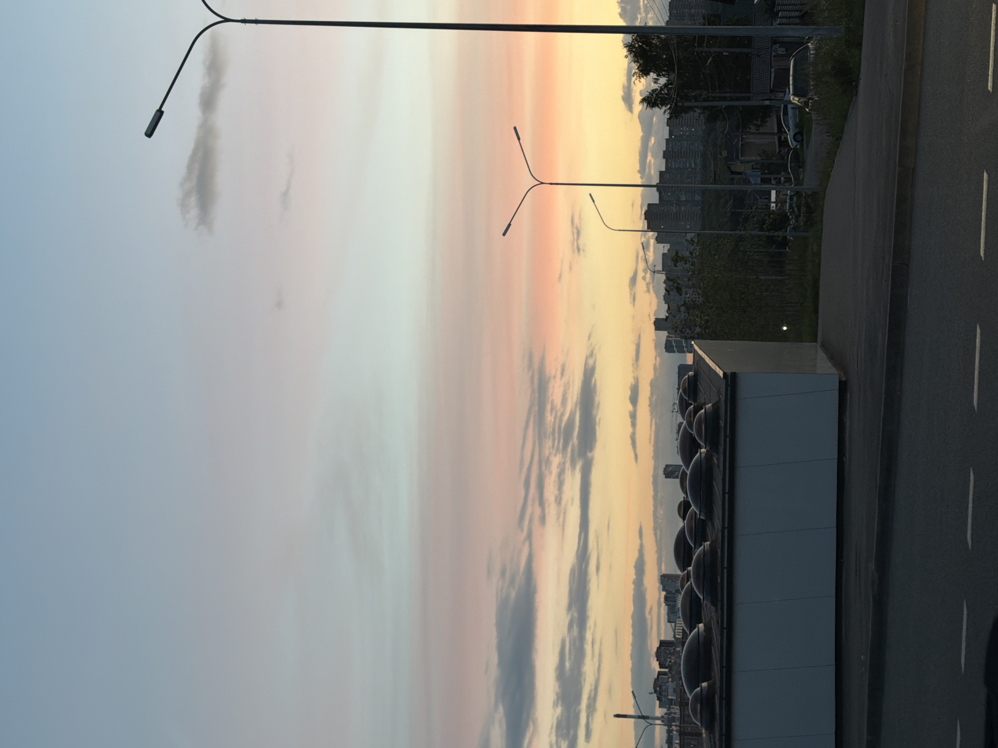 The image depicts a cityscape during a sunset, with a gradient of colors ranging from light blue to pink and orange in the sky. In the foreground, there is a modern, white building with several circular, domed structures on its roof. A paved road runs in front of the building, with white dashed lines indicating lanes. In the mid-ground, several streetlights are visible, along with the silhouettes of apartment buildings in the distance. Trees and greenery can be seen interspersed between the buildings. The scene appears to be an urban or suburban environment. Without further information, the specific location within Kazan, Russia, cannot be determined from the image alone. There are no people or animals visible in the frame.