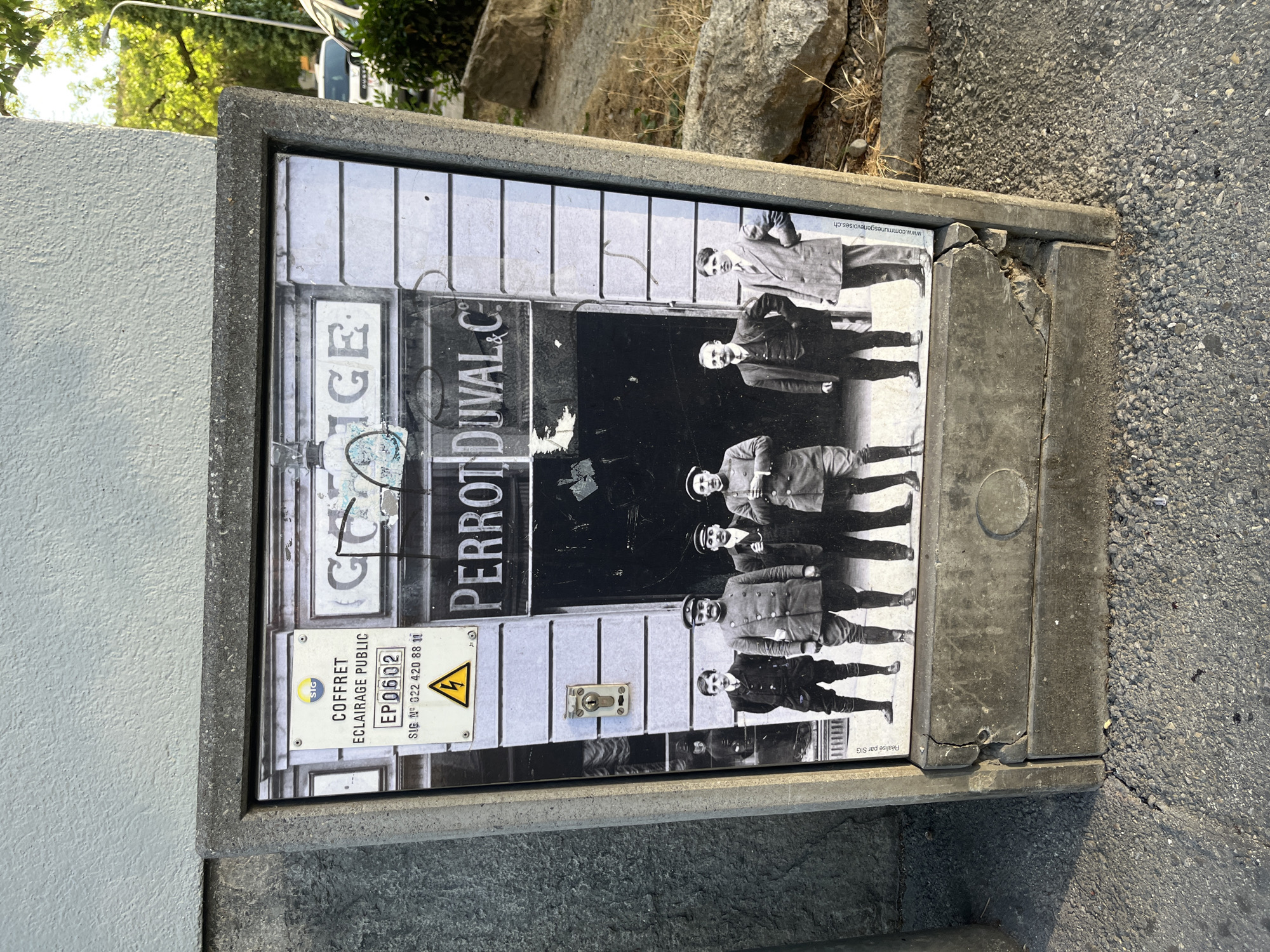 This media file features a photograph on a public utility box, likely located in Geneva, Switzerland. The photograph itself is a vintage black and white image of a group of men, possibly workers, standing in front of a building with "PERROT DUVAL & C" visible on its facade. The men are dressed in formal attire, including suits and hats, suggesting a historical context.
The utility box has a variety of text and symbols. One label reads "COFFRET ECLAIRAGE PUBLIC" with an identification number "EP0602" and a SIG number "022 420 88 11". There is also a yellow triangle with a lightning bolt symbol, indicating danger or high voltage. A website address, "www.communesgenevoises.ch", is also visible, further confirming the Geneva location.
The overall scene appears to be outdoors, with a glimpse of trees and rocks in the background. The lighting suggests it is daytime. The utility box itself shows signs of wear and tear, with cracks and chips in its concrete casing. The photograph on the box is also slightly damaged in some areas. The image has been rotated, so the text and people appear sideways.