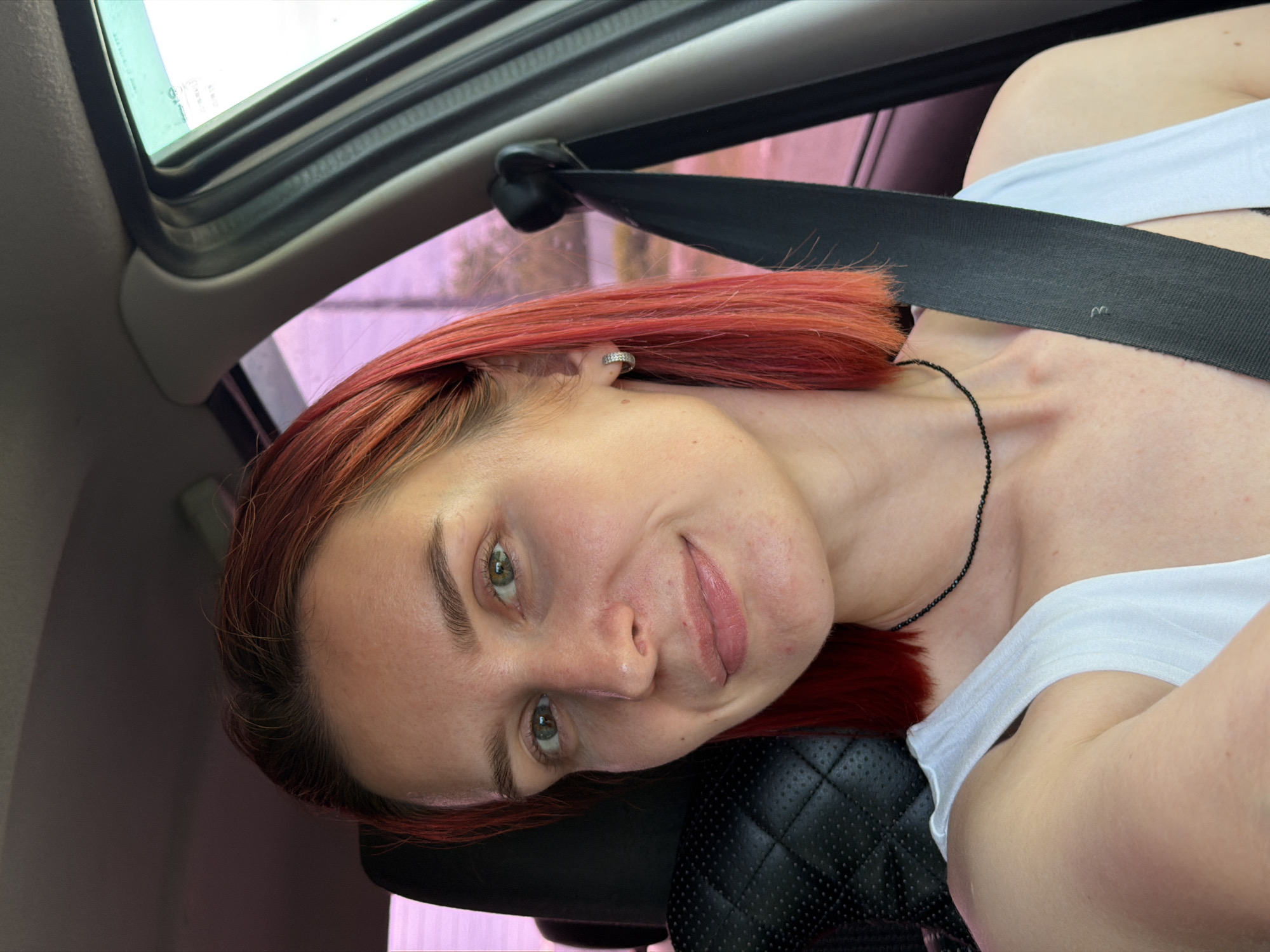 A medium shot depicts a fair-skinned individual with auburn hair sitting in a vehicle, evidenced by the visible seatbelt and sunroof components. The person is wearing a white top and a delicate black necklace. The interior of the car is visible, and the backdrop shows blurred urban elements. While the image does not directly provide information for determining the specific location within the city of Chaikovsky, Russia, it does suggest an urban or suburban setting due to the presence of a vehicle and external building features.
