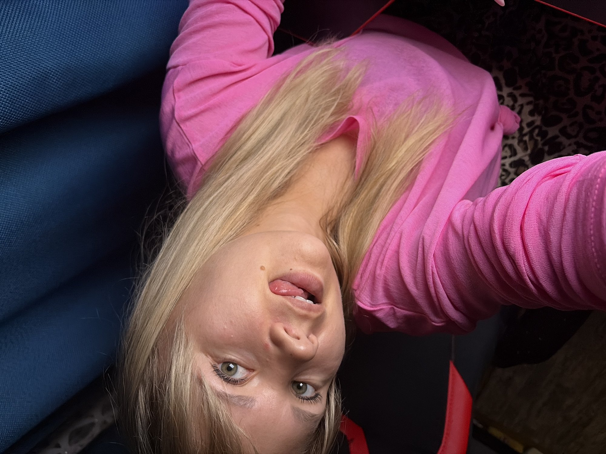 The image is a close-up, slightly angled selfie of a young blonde woman indoors. She is wearing a bright pink long-sleeved shirt. Her head is tilted to her left, and her eyes are open, looking slightly upwards. Her tongue is sticking out from her mouth, touching her upper lip. A small mole is visible on her right cheek. Her hair is straight and falls around her face and shoulders. In the background, a dark blue textured fabric is visible at the top, and a black surface with red accents, possibly a chair, is below her. Leopard-print fabric is visible at the bottom right. The lighting suggests an indoor setting, likely artificial light. There is no clear indication of a specific city or location within Chaikovsky, Russia, as the visible elements are generic to an indoor environment.