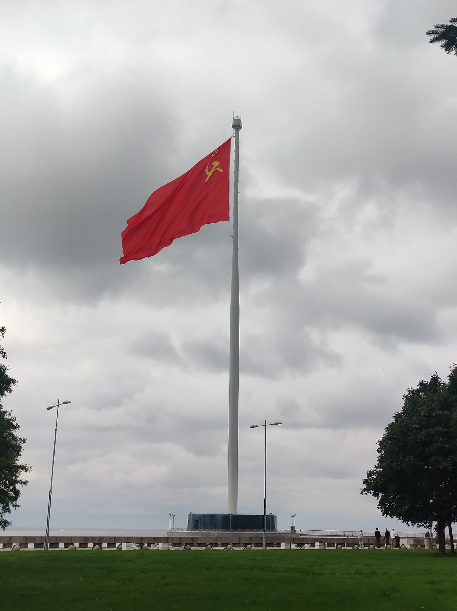 The image shows a large red flag with a yellow hammer and sickle emblem, characteristic of the Soviet Union, flying from a tall flagpole. The flagpole is located on a waterfront promenade. In the background, a body of water, likely the Gulf of Finland, is visible under a cloudy sky. Further along the promenade, there are several people walking and standing, and a modern building with glass panels. Trees and a grassy area are in the foreground, with lampposts lining the promenade. This scene is located at the monument to the heroes of the defense of Leningrad, situated on the Neva River embankment in Saint Petersburg, Russia.