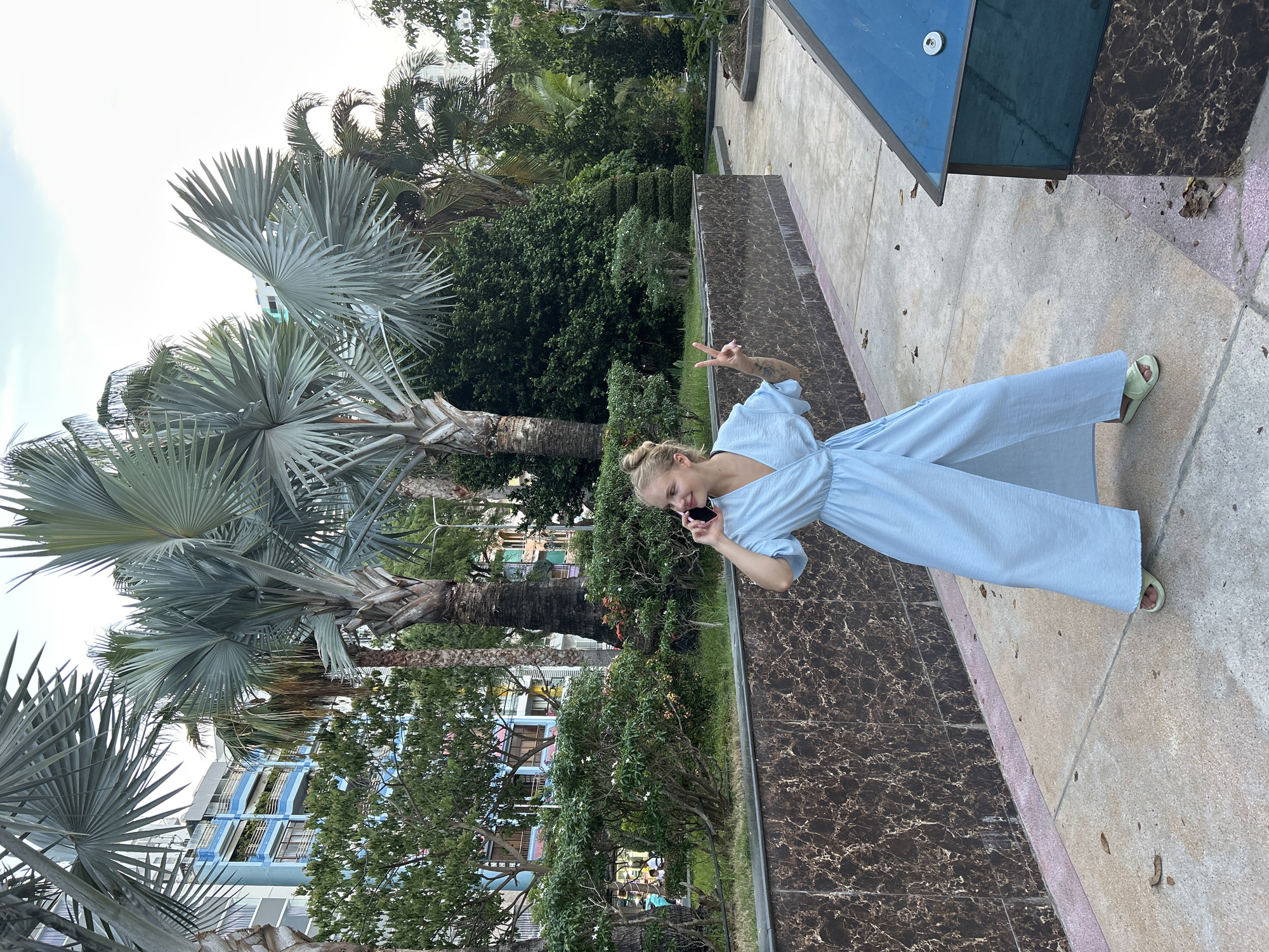 A young woman with blonde hair tied in a bun is standing on a paved walkway in a park-like setting with palm trees and lush greenery. She is wearing a light blue wrap dress and light green slip-on sandals. She is holding a black smartphone to her right ear with her right hand and making a peace sign with her left hand, with her palm facing outward. Behind her, there is a patterned marble wall, a building with blue balconies, and other trees and foliage. The sky is overcast. The location is Nha Trang Ward, Vietnam.