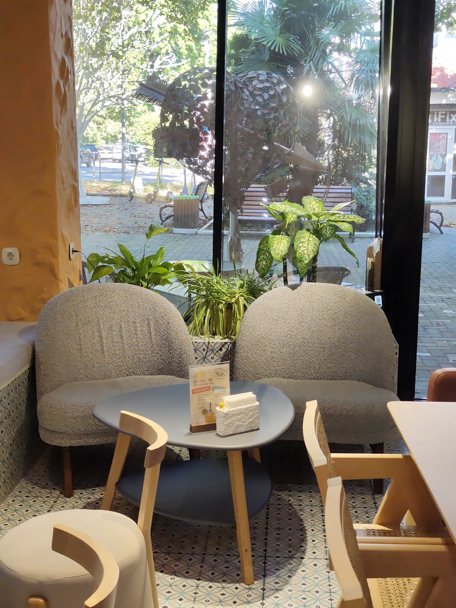 This is an indoor shot of a cafe or restaurant in Sochi, Russia, looking out through a large window to the outdoors. Two round, textured grey armchairs are positioned around a small, round grey table. On the table are napkins and a menu stand. In the foreground, another stool-like seat with a curved wooden element is visible. The floor has a patterned tile design. Outside the window, trees, palm trees, and a large metal sculpture are visible. A bench, trash bin, and electric scooters are also present. The lighting suggests it is daytime, with sunlight filtering through the foliage.