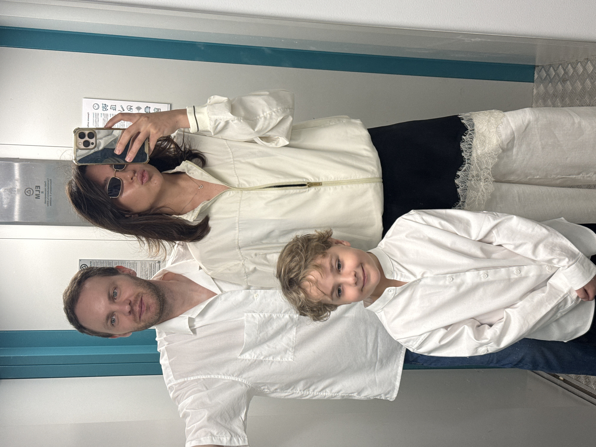 Three people are in an elevator. A woman holds a phone to take a selfie. She wears sunglasses, a white jacket, and a black and white lace skirt. A man in a white button-down shirt stands beside her. A child in a white button-down shirt stands in front of them. The elevator has white walls, a blue stripe, and a metal floor. A sign in Cyrillic script is partially visible. The city of Krasnoyarsk, Russia is not confirmed in this image.