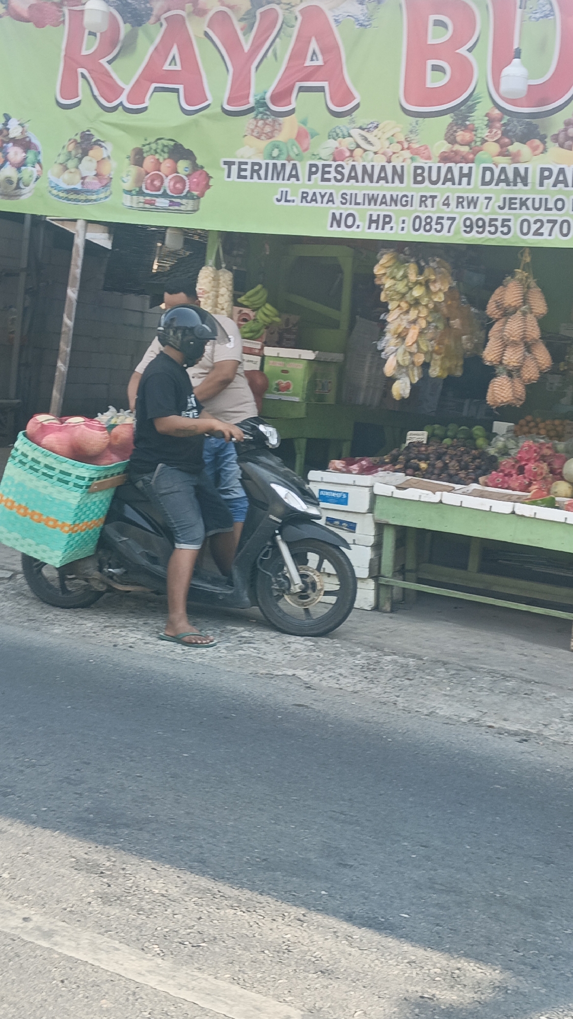 An outdoor scene depicts a fruit stand in Bulungcangkring, Indonesia. A green banner with red lettering, advertising "RAYA BU", hangs above the stall. The banner also displays images of various fruits and text that reads "TERIMA PESANAN BUAH DAN PAN NO. HP: 0857 9955 0270". A man wearing a black t-shirt, denim shorts, and a black helmet is sitting on a black motorcycle with a large turquoise basket filled with red fruits behind him. Another person is partially visible behind him. The motorcycle is parked on an asphalt road in front of the fruit stand, which is laden with a variety of fruits including rambutans, mangosteens, dragon fruits, and bananas. The environment suggests a marketplace setting.