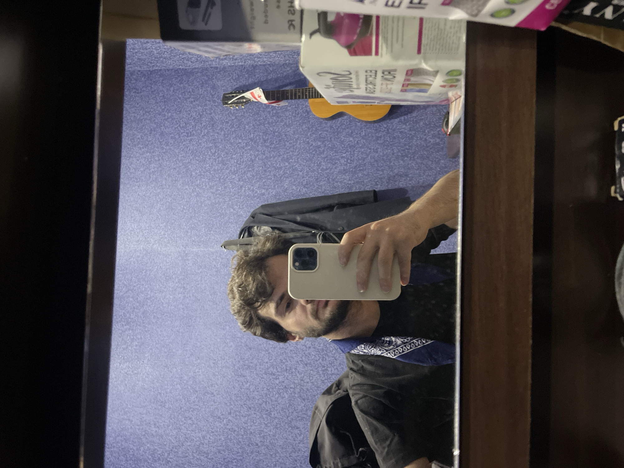 A young man with brown curly hair and a beard takes a mirror selfie in what appears to be a dressing room or backstage area. He is wearing a black t-shirt and a blue bandana with a white paisley pattern around his neck. He is holding a light-colored smartphone in his right hand, with which he is taking the picture. In the background, a dark jacket hangs on a hook on a textured purple wall. To the right, a guitar with a Georgian flag tied to its headstock leans against the wall. A stack of boxes, one of which has the text "Sonos" visible, is to the far right. The reflection in the mirror shows the man, the jacket, the guitar, and the boxes. The foreground of the image shows the top of a wooden dresser or cabinet. The overall lighting is dim. The scene is likely in Tbilisi, Georgia, given the presence of the Georgian flag.