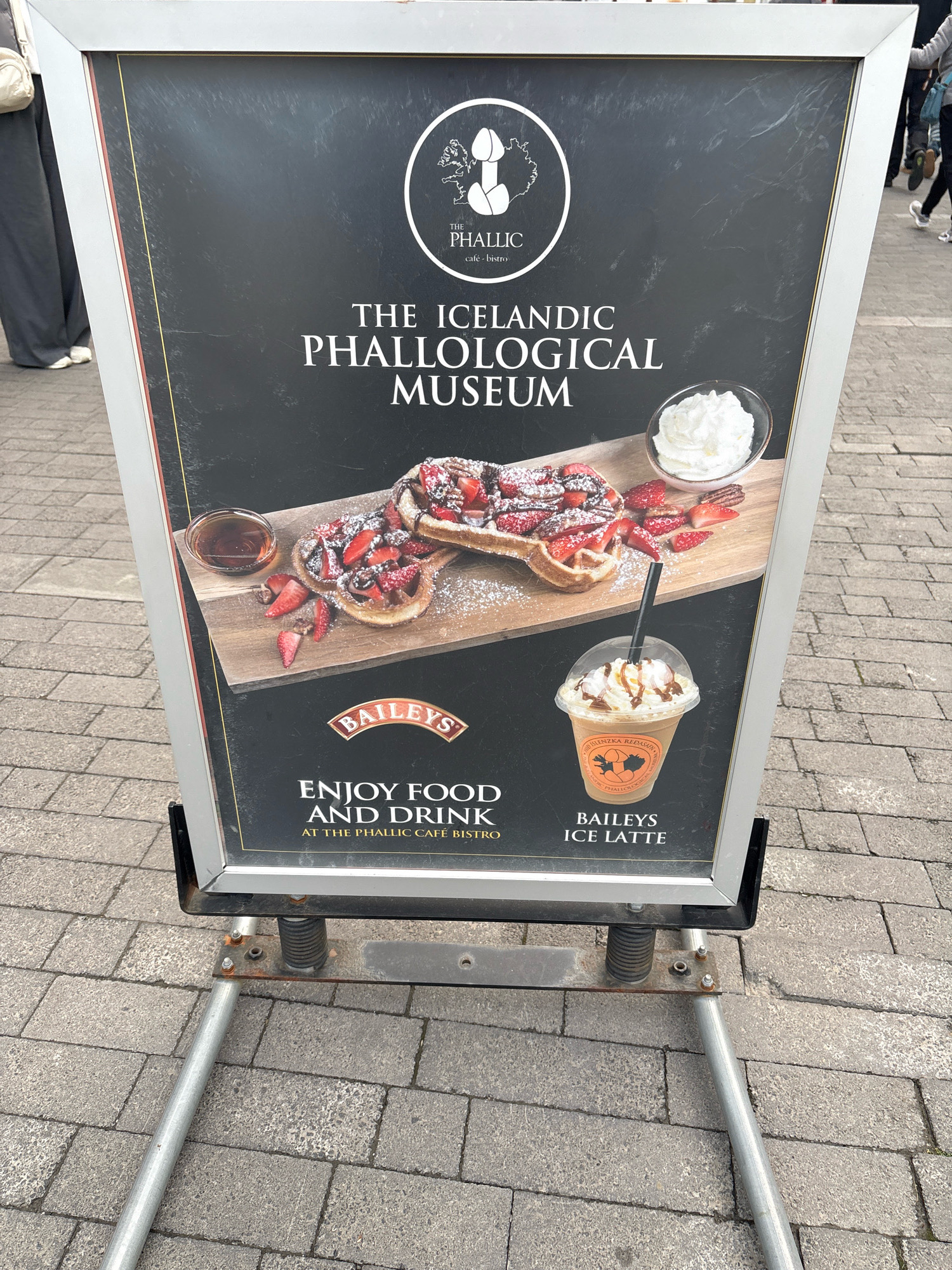 An A-frame sign stands on a paved sidewalk in Reykjavik, Iceland, advertising "The Icelandic Phallological Museum." The sign features a black background with white text and graphics. Prominently displayed is the museum's logo, a stylized depiction of phalluses. Below the logo, the text reads "THE ICELANDIC PHALLOLOGICAL MUSEUM."
The lower half of the sign showcases an advertisement for food and drink. A close-up image displays two waffles topped with strawberries and powdered sugar, accompanied by a small bowl of whipped cream and a glass of syrup. Below this, a Baileys logo is visible, followed by an image of a Baileys Ice Latte in a clear plastic cup. The text "ENJOY FOOD AND DRINK AT THE PHALLIC CAFÉ BISTRO" is also present.
The sign is situated outdoors on a gray, brick-patterned sidewalk. In the background, blurred figures of people can be seen walking, suggesting a public area. The sign's metal frame and supporting legs are visible at the bottom of the image. The overall scene depicts an advertisement for a unique museum and its associated café, highlighting its offerings in a tourist-frequented location.