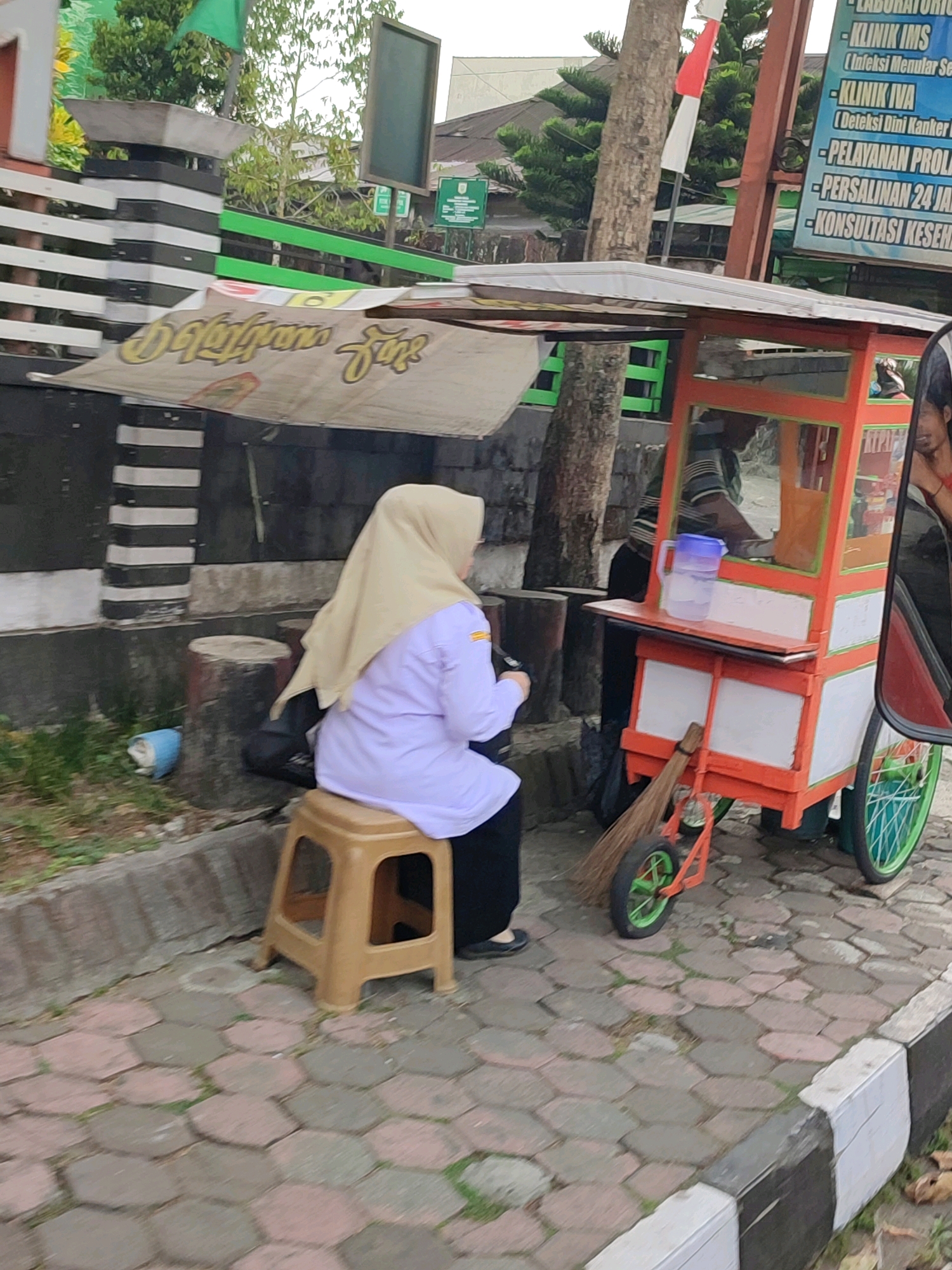A street vendor cart selling drinks is parked on a brick sidewalk in Wonosobo, Indonesia. A woman wearing a beige hijab and a purple shirt sits on a small plastic stool next to the cart. The cart is orange and white with a canopy. A broom is propped against the cart. In the background, a sign with Indonesian text is visible. The scene appears to be on a street with trees and buildings.