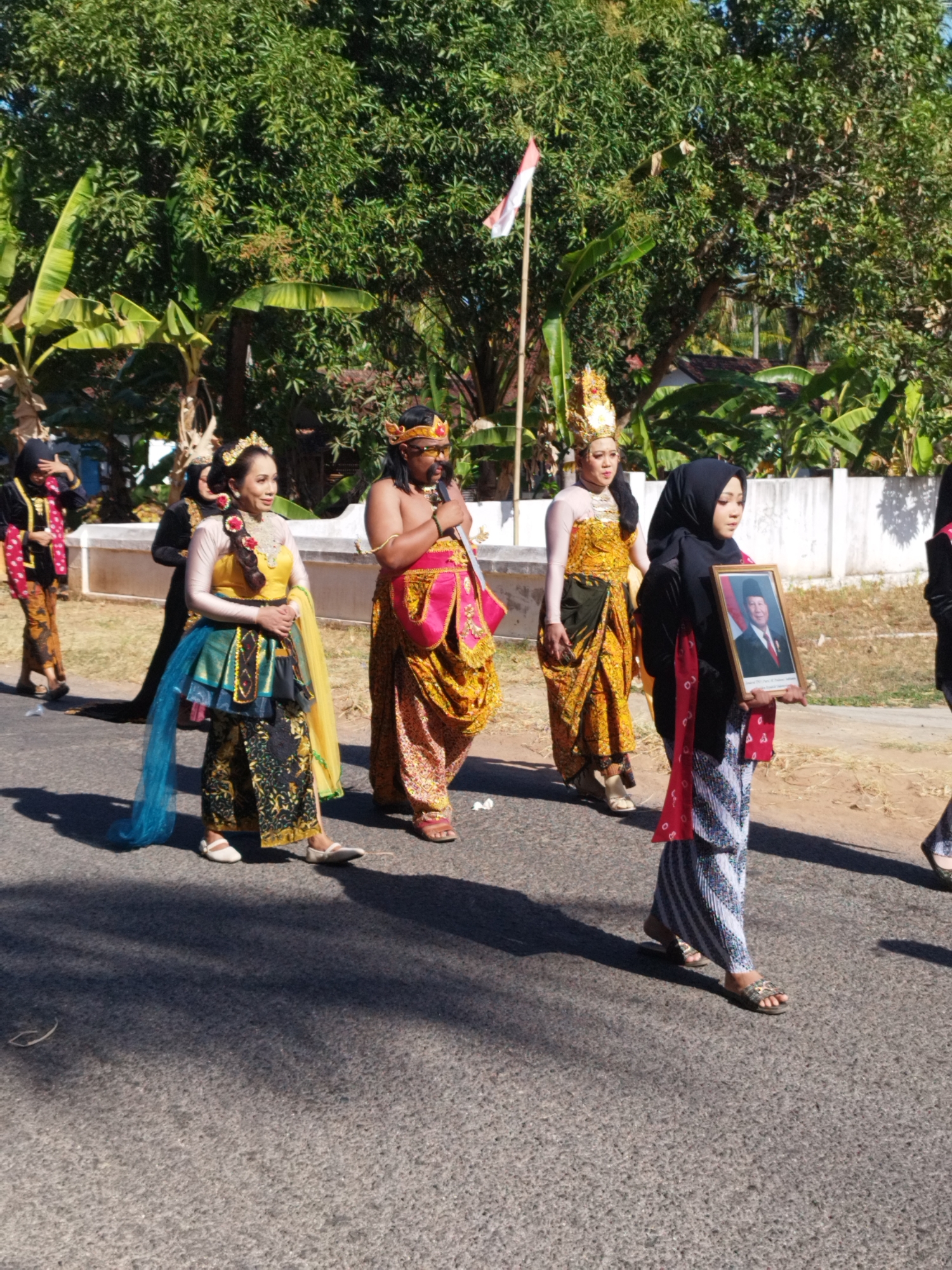 In Andulang, Indonesia, a group of people are walking in a procession. The individuals in the foreground are dressed in traditional attire. A woman in the center wears a yellow top and a layered skirt in blue, green, and black, with a yellow sash. To her right, a man is adorned in a yellow and pink garment, with a golden headpiece. Another woman, on the far right, wears a black hijab and a striped skirt, holding a framed portrait of a man. Behind them, other figures are visible in traditional clothing. The procession is taking place on a paved road, with lush green trees and a white fence lining the background. A flagpole with an Indonesian flag is also present. The scene appears to be under bright daylight.