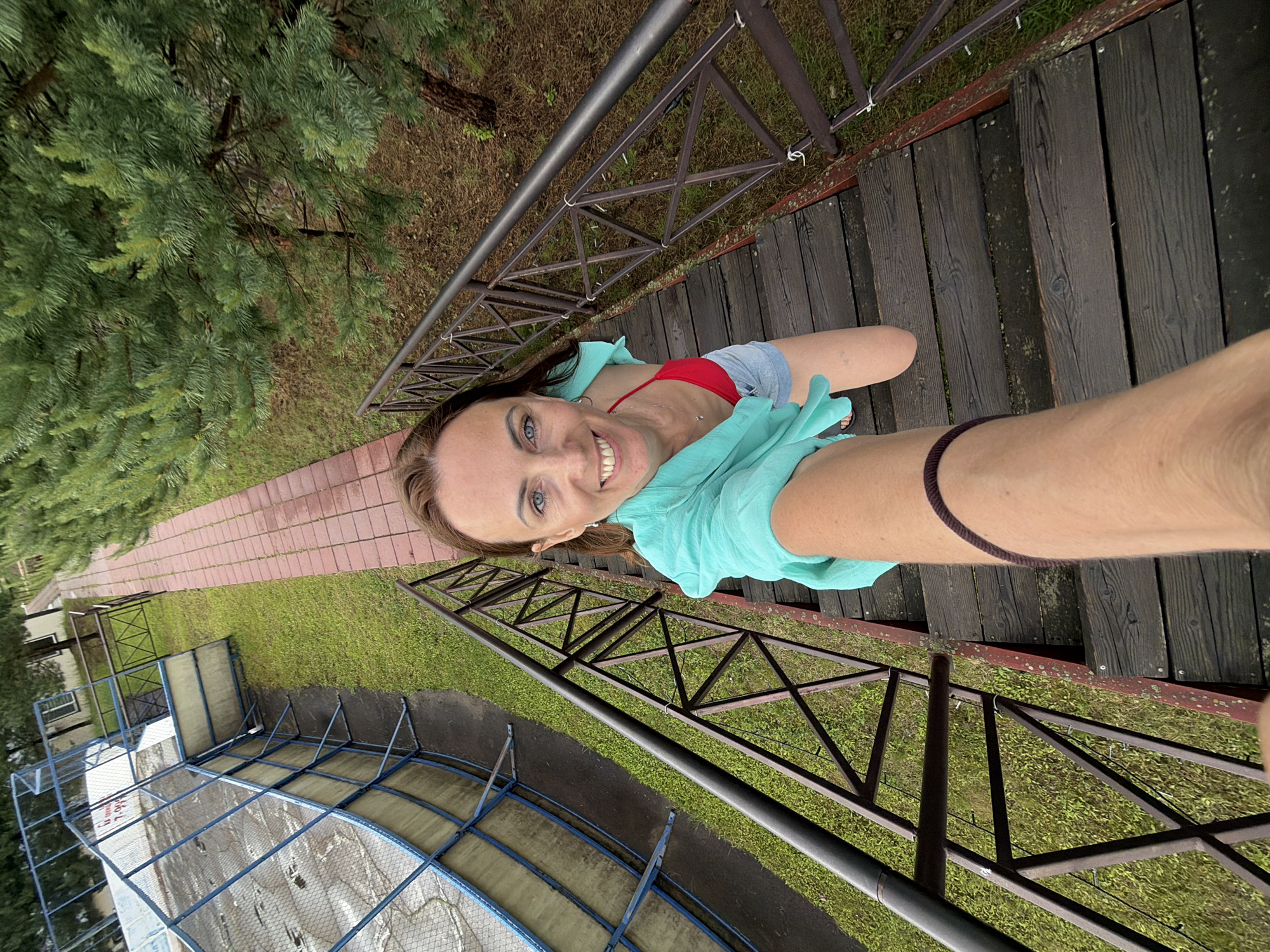 A woman is taking a selfie from a set of wooden stairs outdoors, wearing a red bikini top, denim shorts, and a turquoise cover-up. She is smiling and looking at the camera. The stairs are bordered by a metal railing and are next to a grassy area with some trees. To the left, there is a fenced-off area that appears to be a sports field or court with a blue fence and some netting. The scene suggests a recreational area, possibly a park or sports facility, in Birobidzhan, Russia.