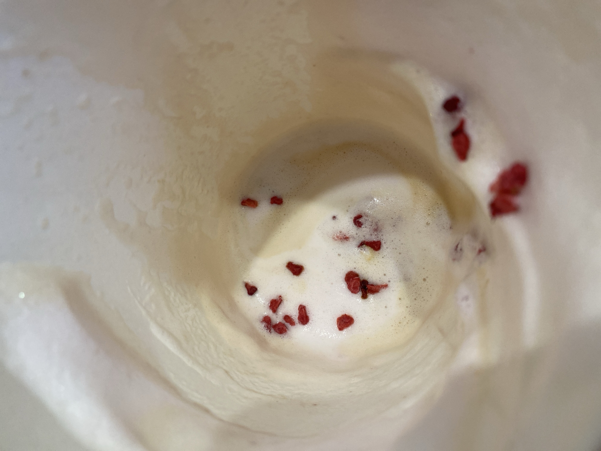 The image shows the interior of a beverage, likely a latte or similar drink, served in a disposable paper cup. The liquid is frothy and light-colored, with several scattered, dried red berries on the surface. The inside of the cup is cream-colored, with a textured surface. There is no indication of the location being Chaikovsky, Russia, nor any people present.