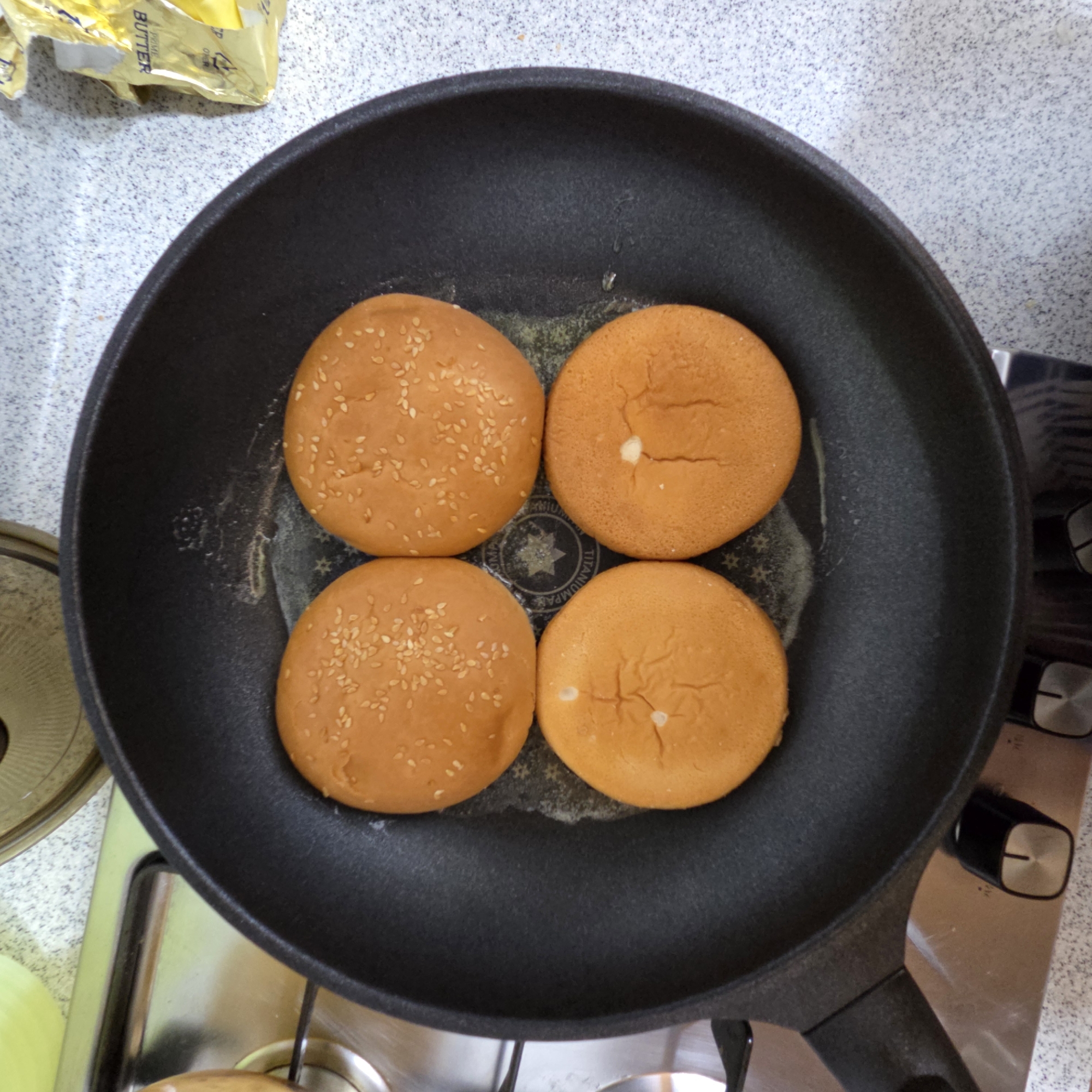 Four burger buns are being toasted in a black frying pan on a stovetop. Two of the buns have sesame seeds on top, and the other two have a white center, possibly from the dough or a filling. The buns appear to be cut in half, with the cut side down in the pan. Melted butter is visible in the pan around the buns. The background shows a speckled countertop and a yellow wrapper, likely for butter. This scene is located in Dunnae-myeon, South Korea.