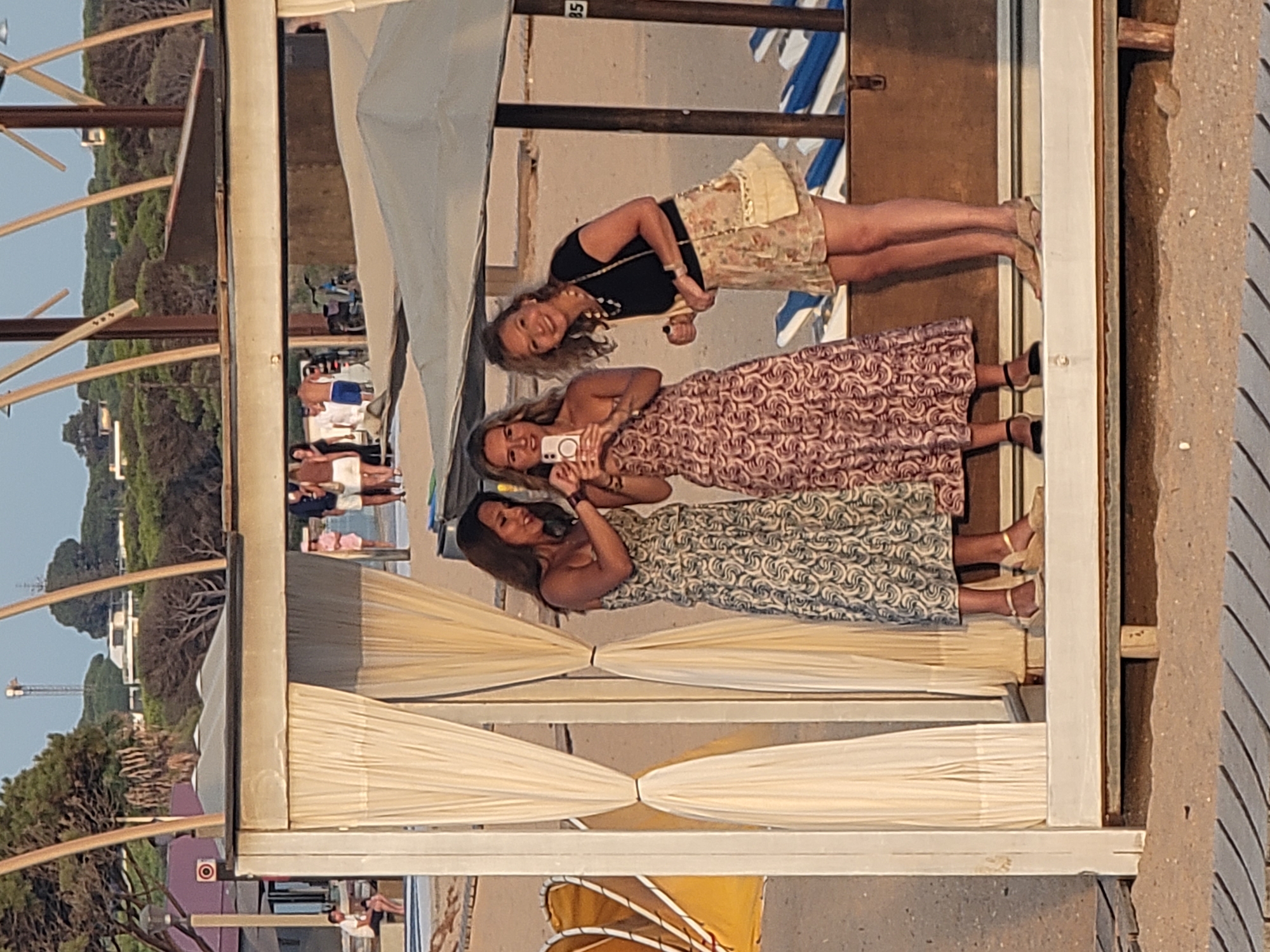 Three women are standing in an outdoor beach cabana in Quarteira, Portugal. The woman on the left is wearing a patterned dress. The woman in the middle is wearing a patterned dress and holding a smartphone. The woman on the right is wearing a black top and a floral skirt. They are all smiling. The cabana has white curtains and wooden posts. In the background, there are beach chairs and trees. The sky is clear and the sun is setting.