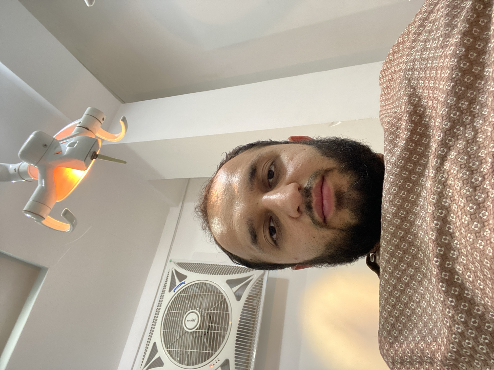 A low-angle, close-up shot shows a man with dark hair and a beard from the chest up, wearing a patterned brown smock. He is in what appears to be a medical setting. Above him, a dental examination light with an orange glow is visible. To his left, a large, white, square fan is mounted on the wall. The background consists of a white wall and ceiling, with some light reflecting off the surfaces. The man is looking directly at the camera with a neutral expression. The environment suggests a dental clinic or similar medical office in Kandahar, Afghanistan, given the fan commonly used in the region and the overall clinic aesthetic.