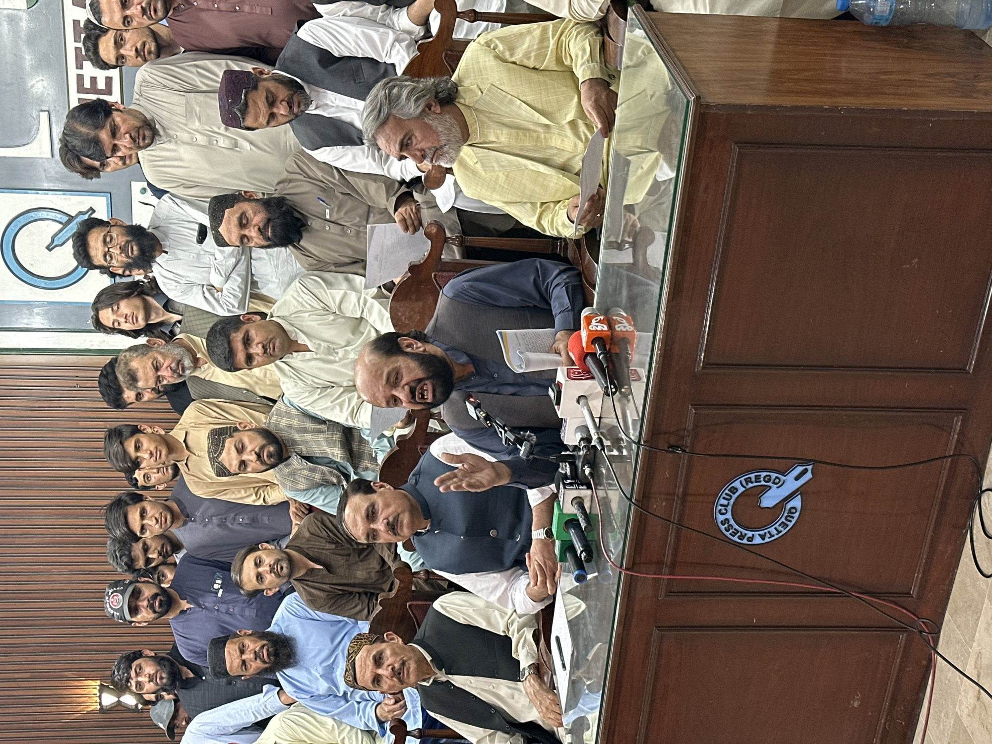 A medium indoor shot depicts a group of men at a press conference in Quetta City Tehsil, Pakistan. Men are seated behind a brown wooden desk with a glass top, some holding papers. Microphones are positioned on the desk. A logo for the Quetta Press Club is visible on the desk. Most of the men are wearing traditional Pakistani attire.