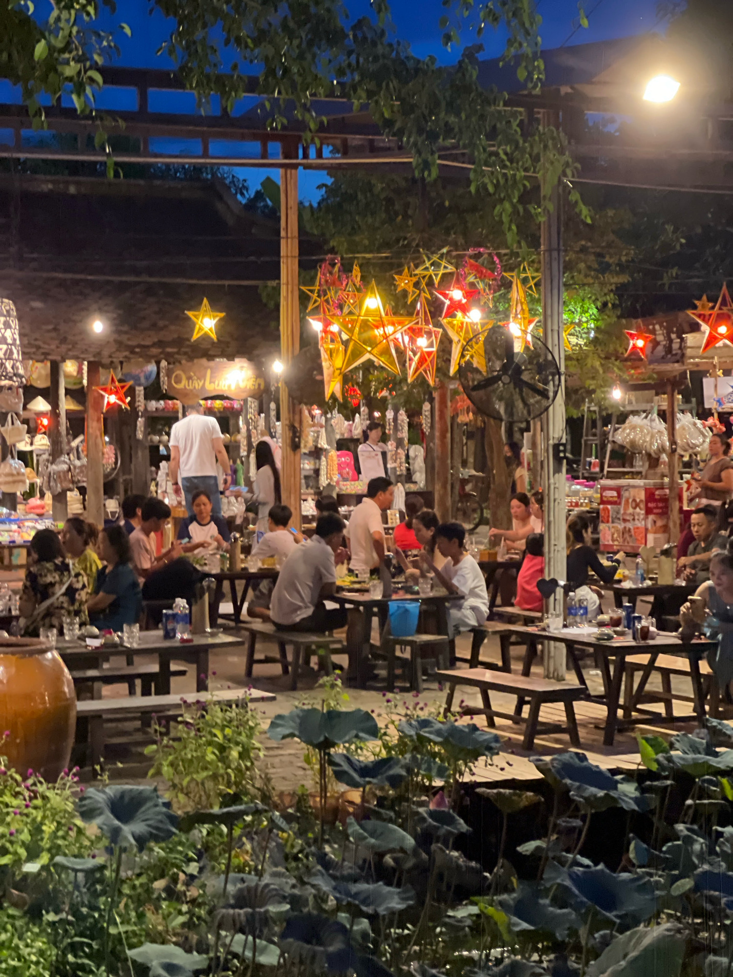 This is a vertical, medium-shot, eye-level, outdoor photograph of a bustling night market in Nha Trang, Vietnam. The scene is illuminated by warm, artificial lights and decorative star-shaped lanterns that hang overhead. In the foreground, lush green water lilies and other plants form a dense border. The midground is filled with people dining at picnic-style tables. The tables are made of dark wood and are laden with food, drinks, and glasses. A large ceramic pot sits to the left of the frame. In the background, stalls display various goods, including hats, bags, and decorative items. Several people are standing and walking around, engaged in conversation and shopping. A ceiling fan is mounted on a pole in the middle ground. The overall atmosphere appears lively and social, characteristic of a popular local gathering spot at night.