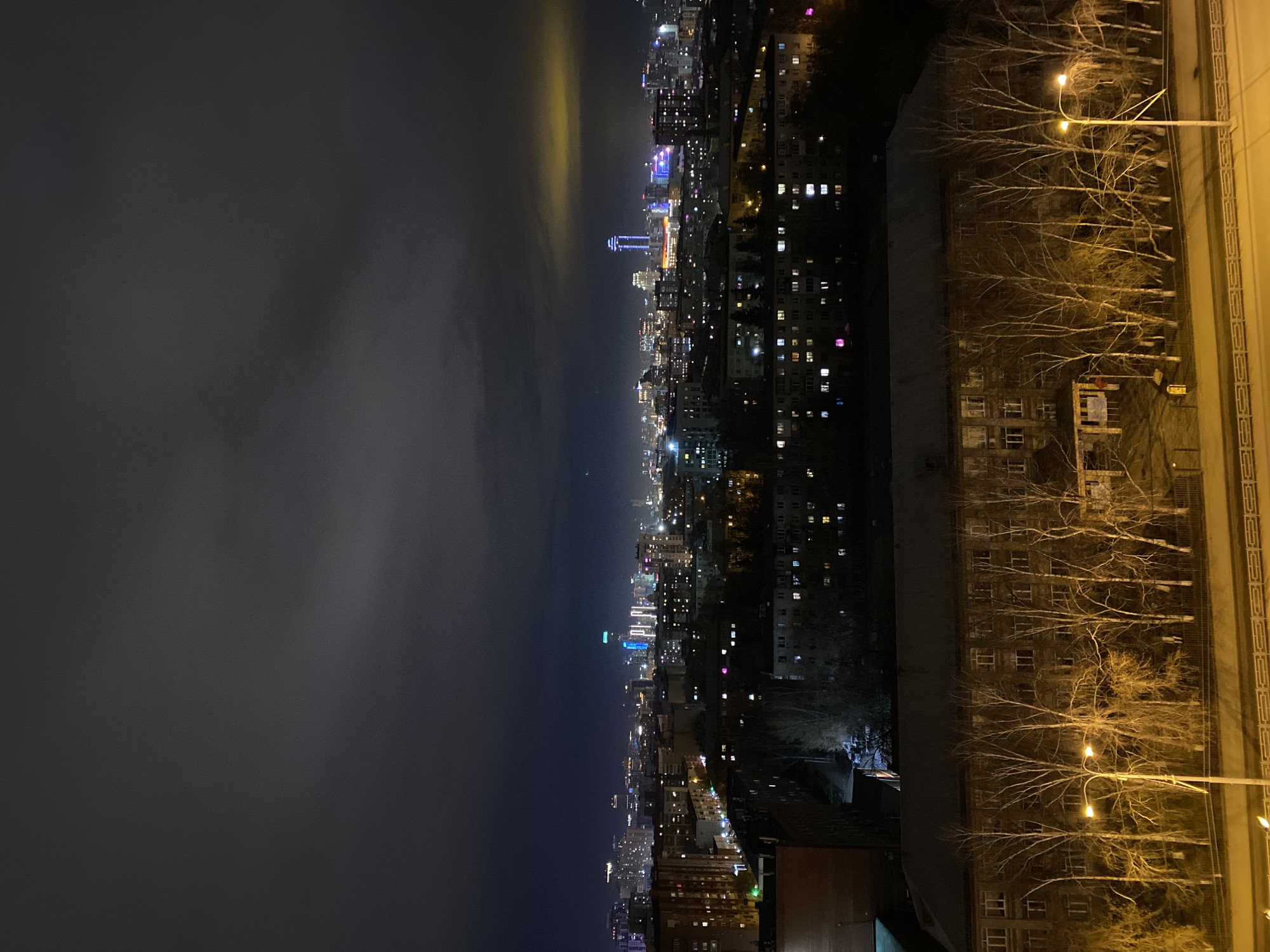 The image shows a nighttime view of Yekaterinburg, Russia, with the city skyline illuminated against a dark, cloudy sky. A prominent tower stands out among the buildings. In the foreground, trees without leaves line a road with streetlights, with residential buildings visible nearby.