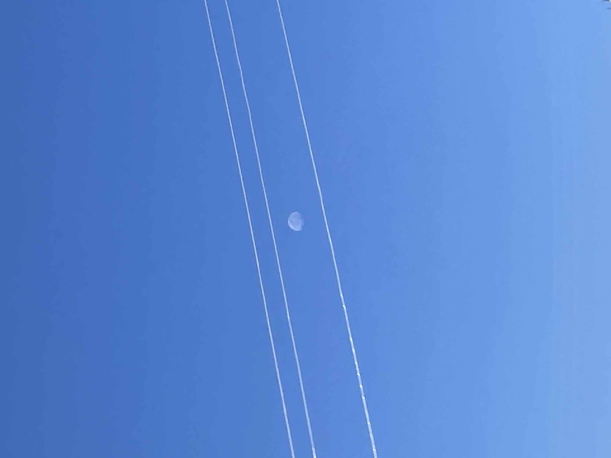 In the city of Avanos, Turkey, three parallel wires, possibly power lines, stretch horizontally across a clear blue sky. Suspended between these wires, near the center of the frame, is a single, small, pale blue object. Its shape is indistinct, appearing somewhat circular or oval, and it seems to be softly illuminated, suggesting it might be a balloon or some other airborne object. The rest of the image is dominated by the vast expanse of the sky, devoid of any other visible elements.