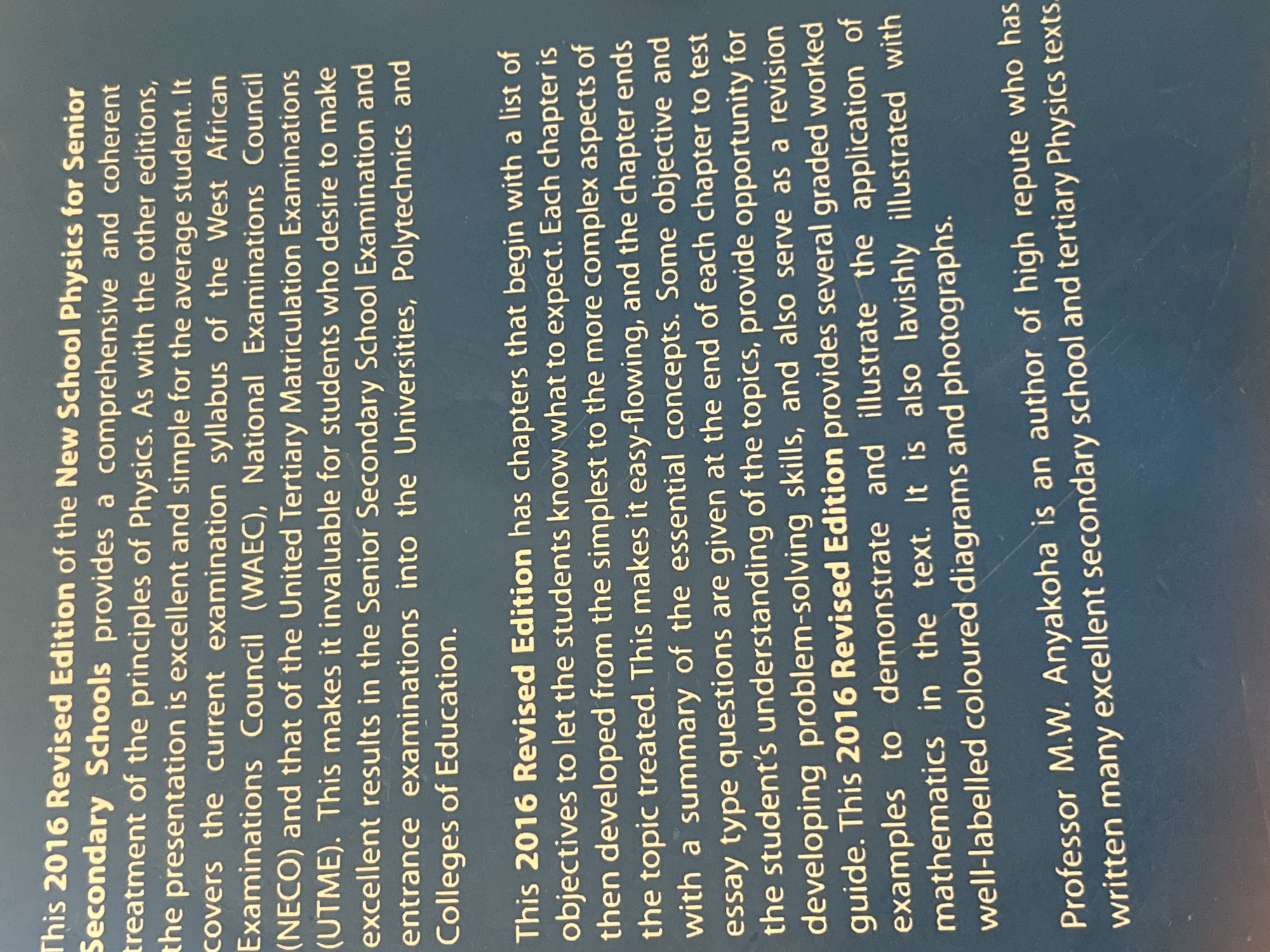 This image is a close-up, angled shot of a book cover or dust jacket, with the text oriented vertically. The background is a dark, textured blue. The primary content is the book's blurb, describing a physics textbook.
The text identifies the book as the "2016 Revised Edition of the New School Physics for Senior Secondary Schools." It emphasizes a "comprehensive and coherent treatment of the principles of Physics" that is "excellent and simple for the average student." The book is stated to cover the syllabi of the West African Examinations Council (WAEC), National Examinations Council (NECO), and the United Tertiary Matriculation Examinations (UTME), making it valuable for students aiming for excellent results in secondary school and for entrance examinations into higher education.
The blurb also highlights features of the 2016 Revised Edition, including chapters starting with objectives, a progression from simple to complex topics, summaries of concepts, and objective and essay questions at the end of each chapter. It also mentions several graded worked examples to illustrate mathematical applications and notes that the book is "lavishly illustrated with well-labelled coloured diagrams and photographs."
The author is identified as Professor M.W. Anyakoha, described as an "author of high repute who has written many excellent secondary school and tertiary Physics texts."
The image itself does not depict any people, objects other than the book's text, or a specific scene. The setting is implied to be a publication or retail environment where this book is presented. There are no discernible details about time of day, weather, or emotions. The location context provided, Jauro Yinu, Nigeria, suggests the origin or intended audience of the textbook, particularly given the mention of Nigerian examination bodies.