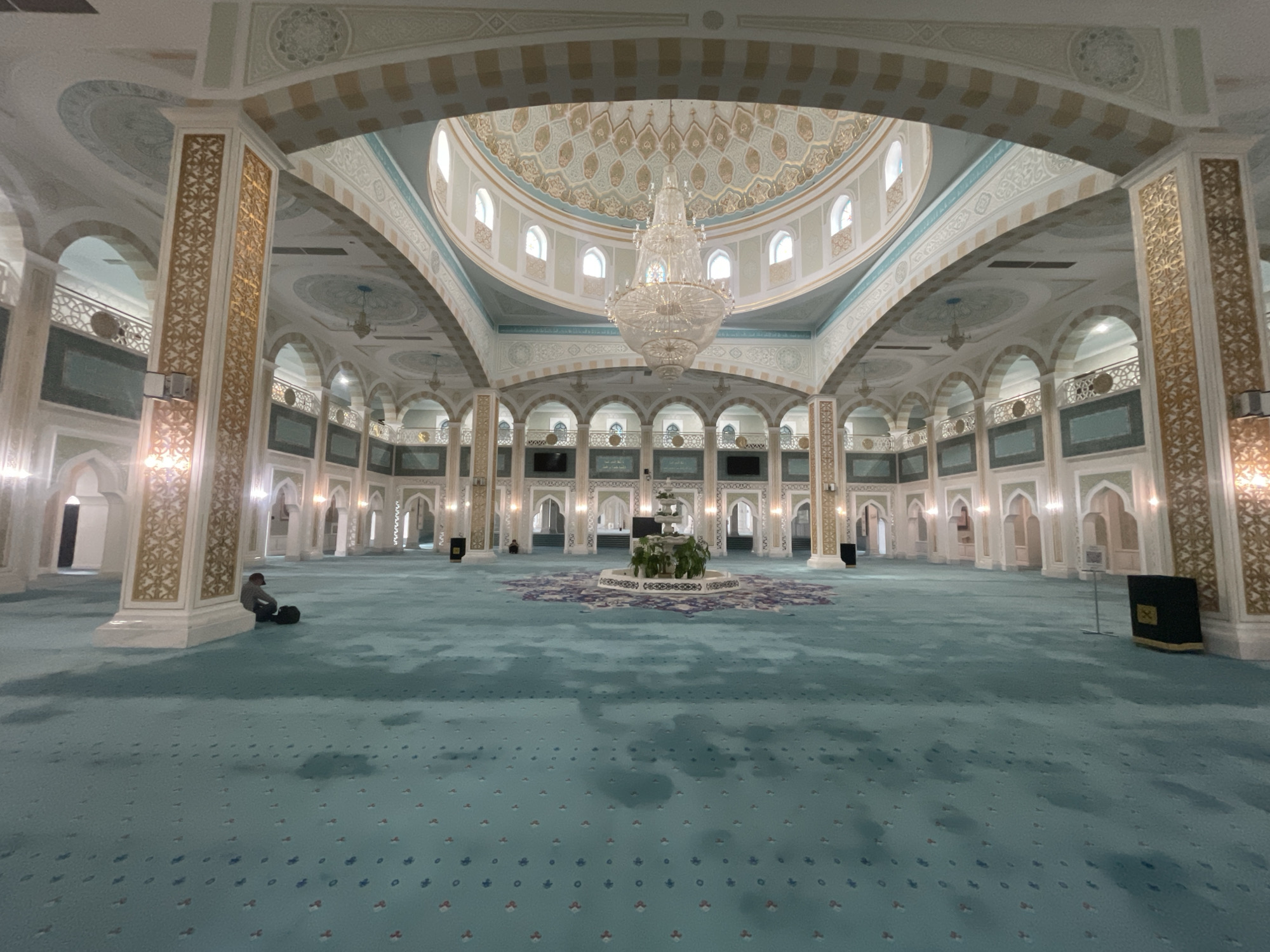 The image depicts the interior of the Hazrat Sultan Mosque in Astana, Kazakhstan. The expansive prayer hall is covered in a blue patterned carpet. A man is sitting on the floor near a pillar on the left side of the frame. A fountain is in the center of the room, surrounded by a floral arrangement. The mosque's architecture includes large arches, pillars with golden ornamentation, and a central dome with a chandelier. Rows of arches and pillars extend into the distance.