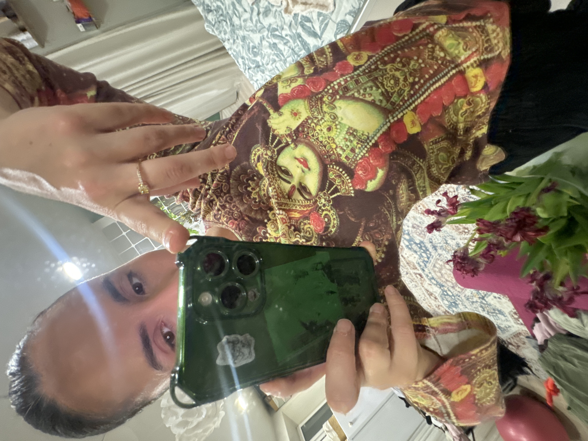 A person is taking a selfie with a phone in a green case. The person has dark hair and is wearing a brown patterned top with an image of a woman on it. The person is wearing a ring on their left hand. The background includes a bed with patterned bedding, a bouquet of flowers, and some clothes. It is not possible to identify the location in Krasnoyarsk, Russia based on the information given.
