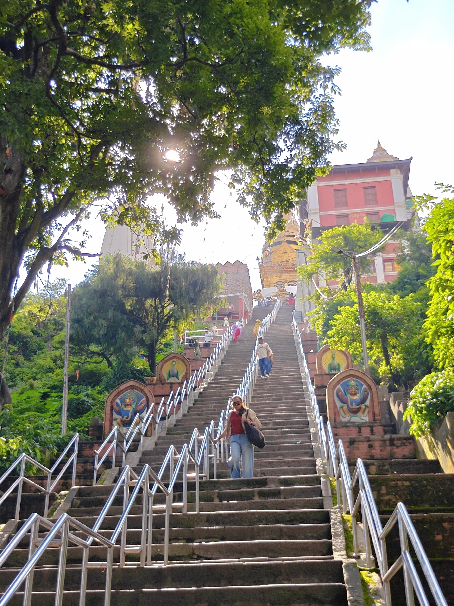 This image captures a wide stone staircase leading upwards towards a golden Buddhist stupa, prominently visible in the background. The staircase is flanked by decorative archways featuring colorful paintings of mythical creatures. Lush green trees and foliage surround the stairs and buildings, with sunlight filtering through the leaves. Several individuals are seen ascending the stairs. In the foreground, a woman with sunglasses and a beige jacket is walking down, carrying a dark bag. Further up, a man in a light-colored shirt and blue jeans is walking upwards. The architectural style suggests a location in Kathmandu Metropolitan City, Nepal, likely a temple complex or sacred site, given the presence of the stupa and the religious iconography. The overall scene depicts a sunny day with moderate foot traffic on the sacred steps.