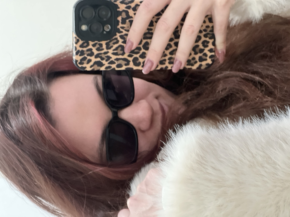 The scene, set in the city of Budva, Montenegro, features a female subject captured in a close-up, likely a selfie. Her head and upper torso are visible, with long, wavy reddish-brown hair framing her face. She wears dark, rectangular sunglasses that obscure her eyes. A subtle smile is discernible on her mouth. A prominent white, fluffy, or fur-lined garment covers her left side and shoulder. Her right hand, featuring manicured nails with a light pink or nude polish, holds a smartphone in front of her. The smartphone is encased in a distinct leopard print cover, and its rear camera module, equipped with multiple lenses and a flash, is clearly visible. The background is a plain, indistinct light-colored surface, suggesting an indoor environment.