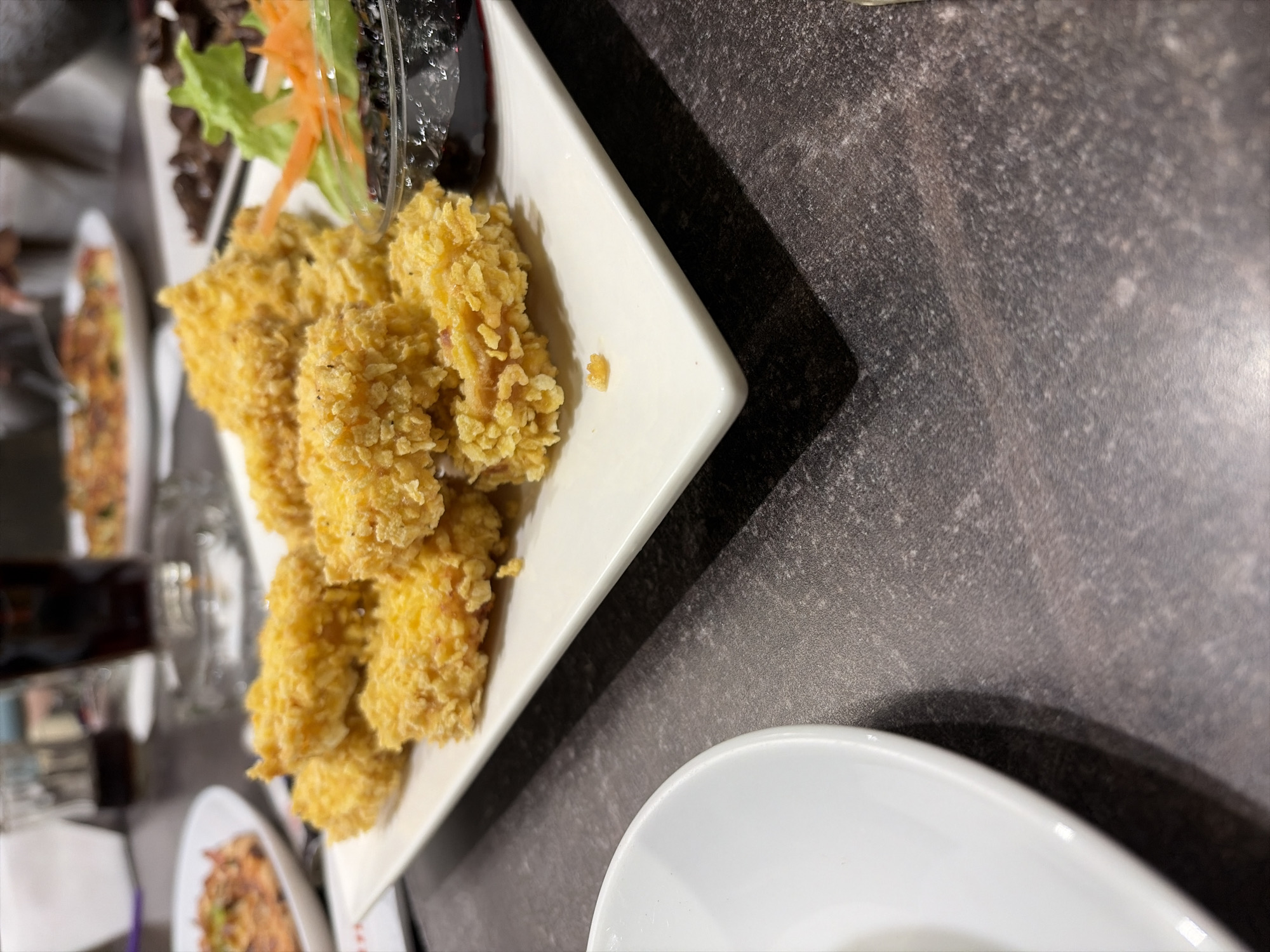 The image shows a table setting with a variety of dishes. In the foreground, there is a white plate with fried, breaded food, possibly chicken or fish. The food is golden brown and appears to be crispy. Next to it, a white bowl is partially visible in the lower right corner. In the background, there are other plates with various foods, including salads and possibly other appetizers. The table has a gray, mottled surface. The location is Unknown, Unknown.