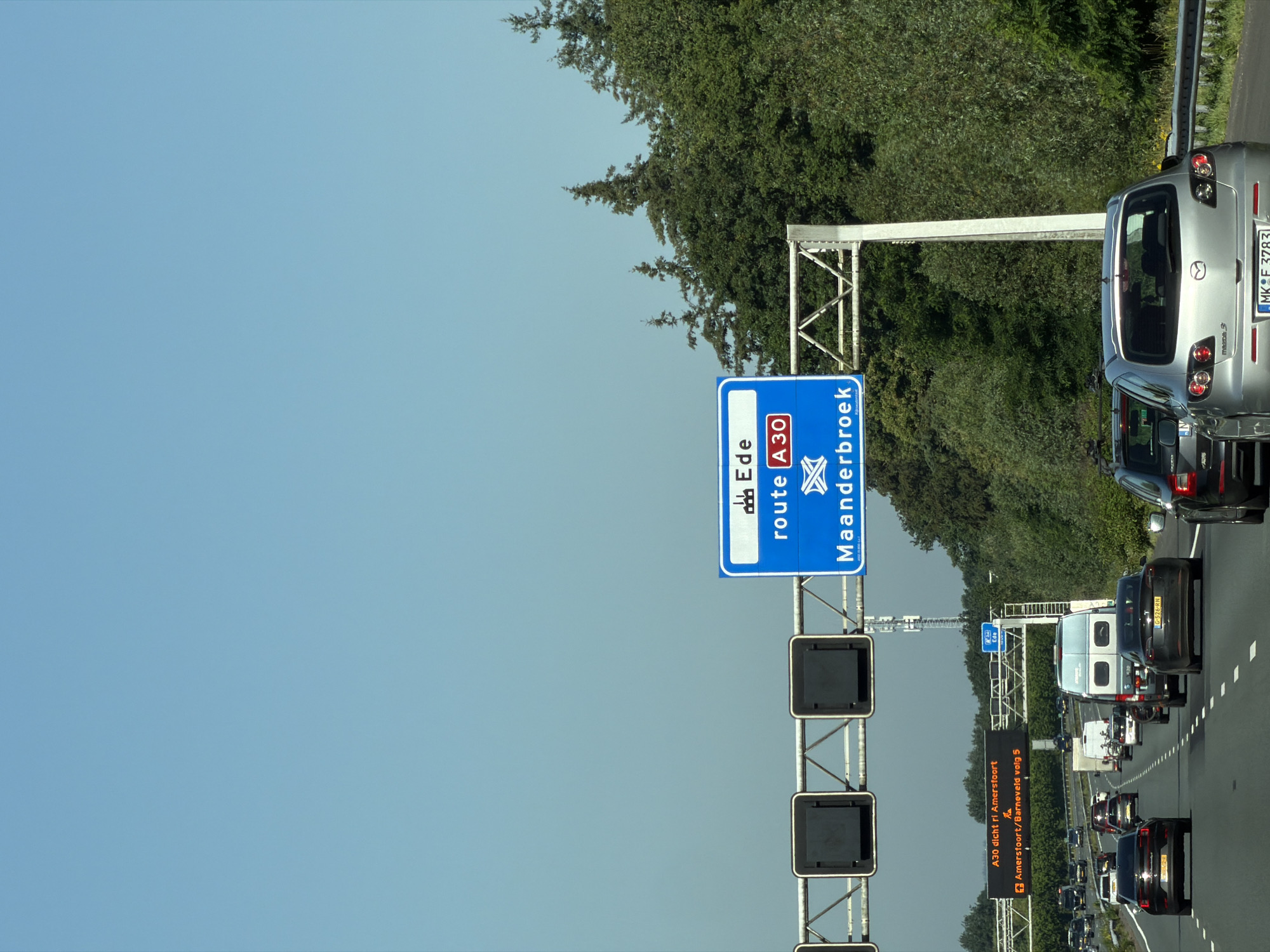 The image shows a highway scene with several cars and signs. A large blue overhead sign indicates "Ede" and "route A30 Maanderbroek." To the left, another electronic sign displays "A30 dicht ri Amersfoort" and "Amersfoort/Barneveld volg 5." Several vehicles are visible on the multi-lane highway, including sedans, a van, and a camper. The environment is characterized by a clear blue sky and lush green trees lining the side of the road. The overall impression is a typical highway landscape in the Netherlands, likely near Bennekom, given the mention of Ede and Amersfoort.