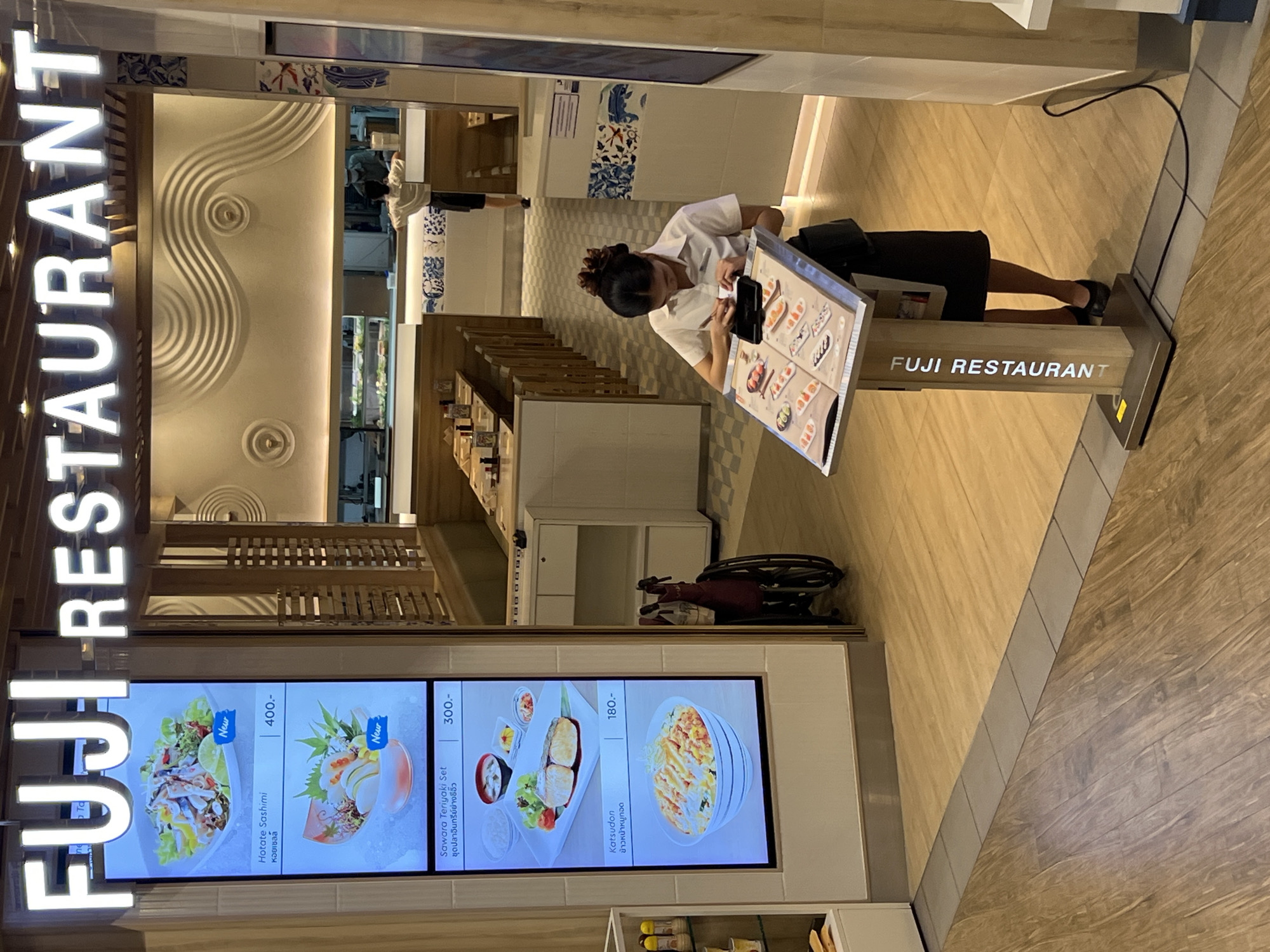 The image depicts the exterior entrance of a Fuji Restaurant, a Japanese dining establishment, situated in Pattaya City, Thailand. Prominently displayed above the entrance is the restaurant's name in large, illuminated letters. To the left, a digital menu board showcases various Japanese dishes, including "Hotate Sashimi" and "Sawara Teriyaki Set," with their respective prices. Inside the restaurant, customers are seated at wooden tables, and behind the counter, staff can be seen preparing food. A woman, dressed in a white uniform and black skirt, is standing by a display stand, perusing a menu. In the foreground, a wheelchair is positioned near the entrance, suggesting accessibility for disabled patrons. The flooring is a mix of polished wood and tiled surfaces, creating a modern and clean aesthetic. The overall ambiance suggests a well-maintained and inviting dining space.