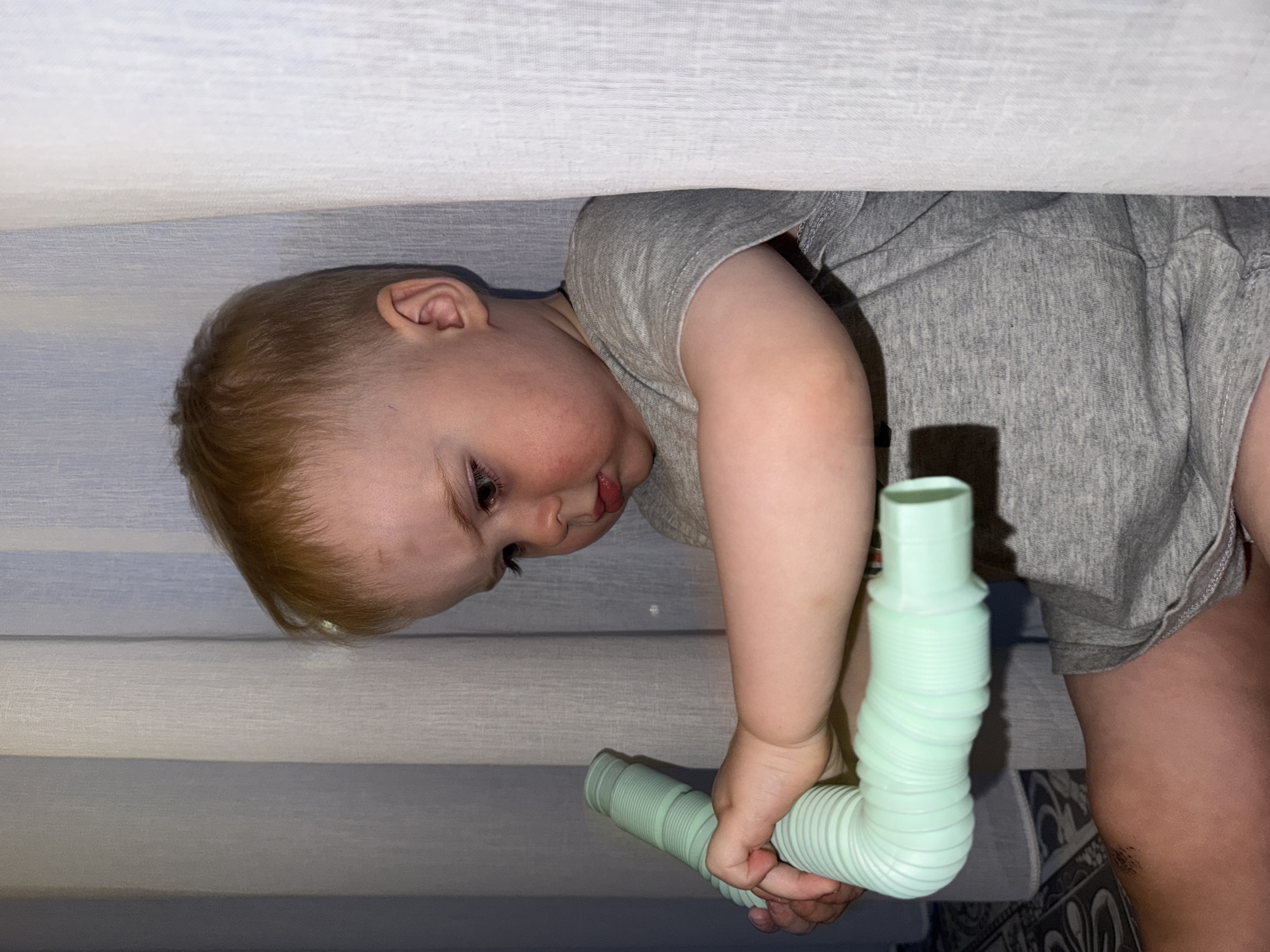 A young child with fair skin and reddish-blonde hair is partially visible under a white or light-colored surface (possibly a table or bed) with horizontal linear patterns. The child is wearing a gray short-sleeved shirt and is holding a light green, corrugated plastic tube in their hands. The child is looking down at the tube. Part of an adult leg is visible in the lower right of the frame. The background is blurry. There is no information to indicate this image was taken in Votkinsk, Russia.