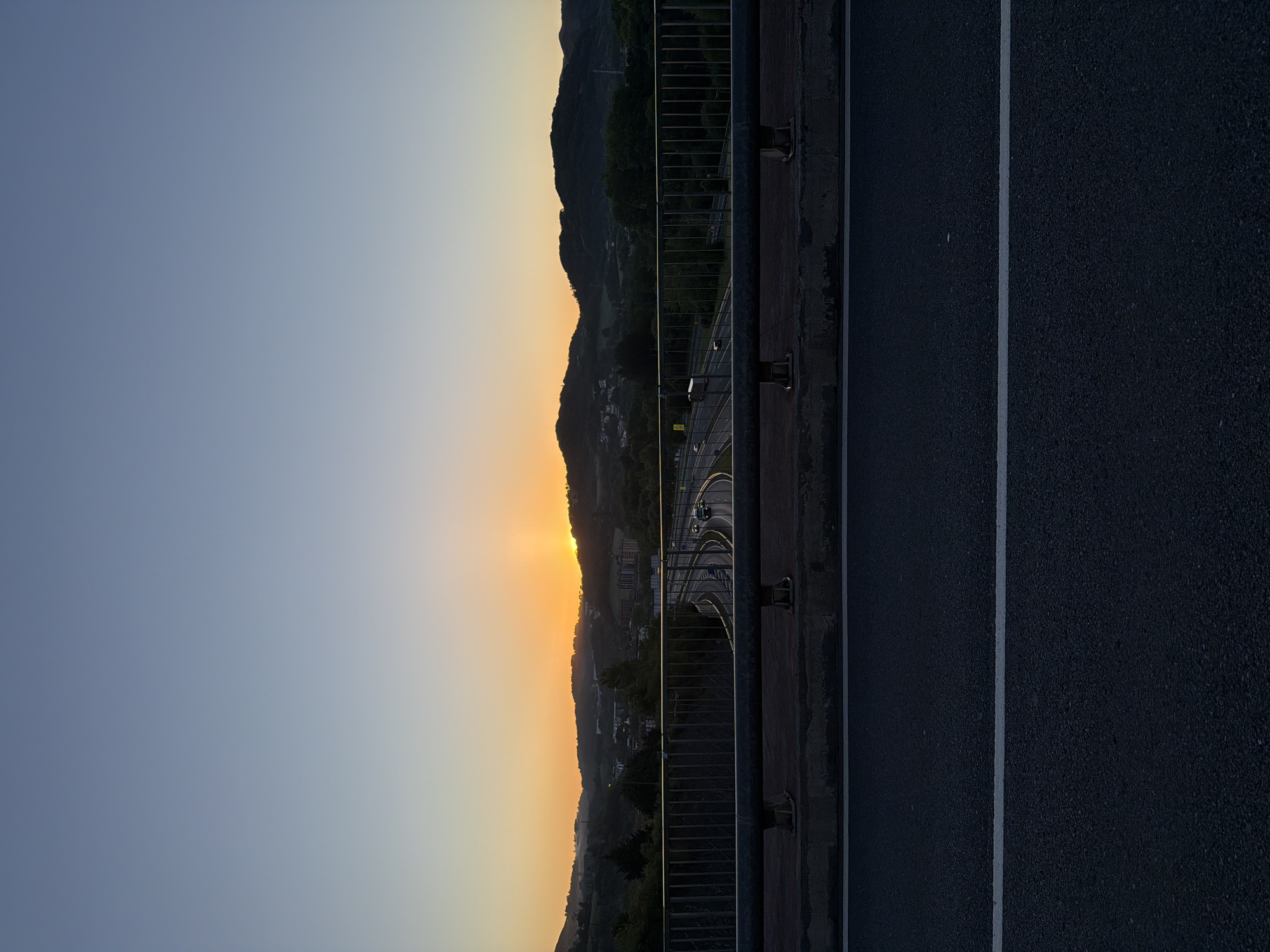 The image captures a sunset view from a bridge in Errenteria, Spain. The sun is setting over the hills in the background, creating an orange glow. The bridge has a metal railing, and on the right side of the image, the road surface is visible with a white line marking the lane. There are cars on the highway below. The sky is a gradient of blue and orange. The image is taken from a slightly elevated perspective.