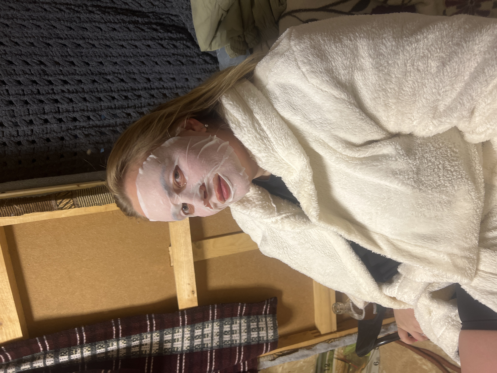 A woman is wearing a white terrycloth robe and a sheet face mask. She is indoors, lying on a bed with a dark gray textured blanket and a green jacket. Behind her is a light brown canvas frame resting against a wall decorated with a maroon, tan, and white patterned textile. The location in Chaikovsky, Russia cannot be determined from the image.