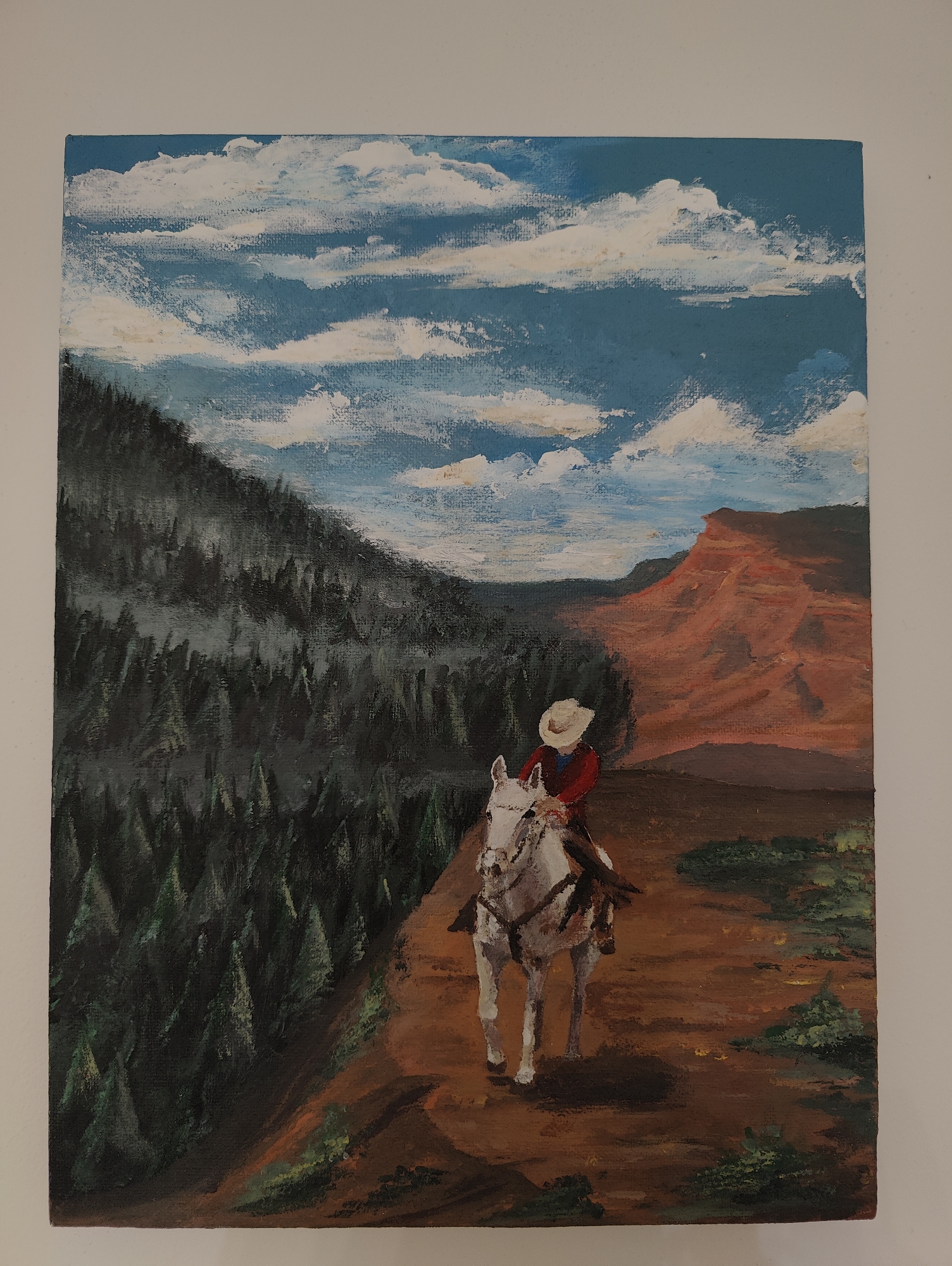 A rectangular canvas painting depicts a person on horseback riding on a trail. The rider, wearing a red shirt and a white hat, is facing away from the viewer. The horse is white with brown markings. The trail is brown and appears to be along the side of a mountain or hill. To the left, a dense forest of dark green trees covers the mountainside. In the background, a large red rock formation is visible, and above it is a blue sky with white clouds. The overall style of the painting suggests it is an amateur work, with visible brushstrokes and somewhat simplistic rendering. The painting's location is not identifiable as being in Ipswich, United Kingdom, from visual cues alone.