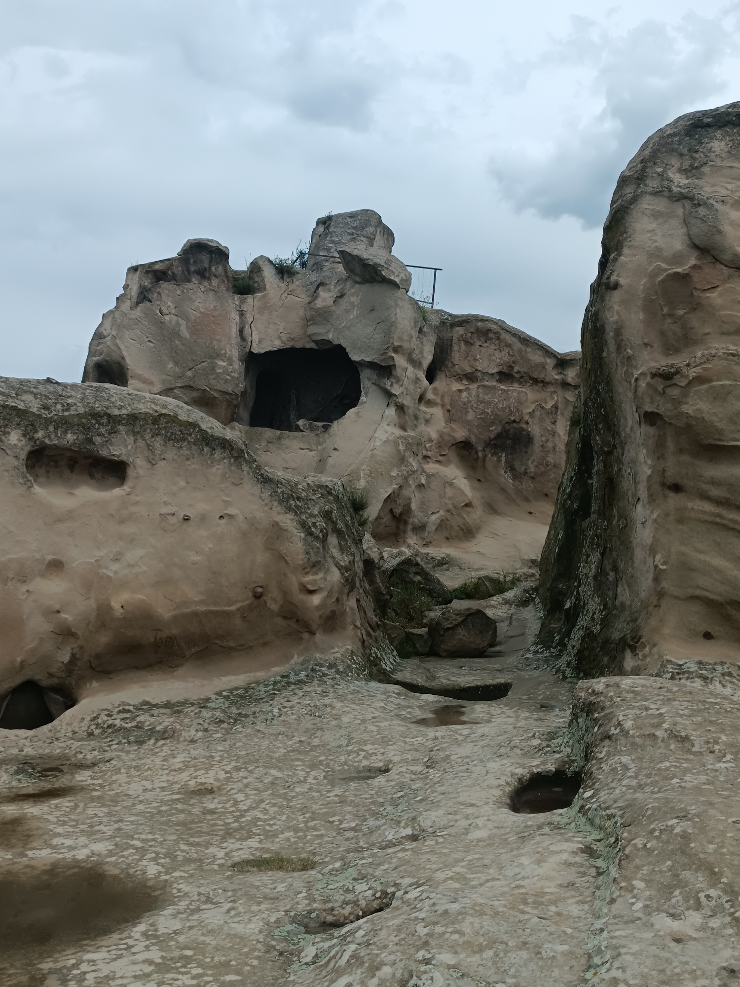 The image shows a view of the ancient cave city of Uplistsikhe, Georgia. The foreground features a rough, uneven stone surface with several indentations, some of which contain water. To the left, a large, weathered rock formation rises, displaying natural erosion patterns and a dark opening that suggests a cave or structure. Further into the scene, a narrow passage leads between imposing rock faces. The right side of the image is dominated by a tall, textured rock formation with streaks of green lichen. In the background, a larger rock massif with a prominent cave entrance is visible, topped by a dark opening. A small metal structure, possibly a railing or fence, is partially visible on top of the rock formation to the left. The sky above is overcast with gray clouds. The overall impression is one of a natural, ancient, and somewhat rugged landscape, hinting at historical human habitation within the rock.